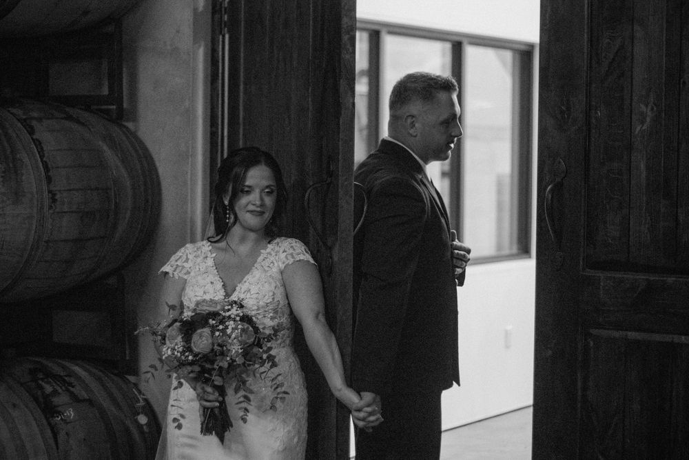 Black and white photo of bride holding bouquet and groom holding hands at winery wedding venue with wooden barrels.