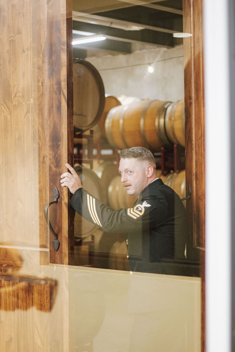 Military officer in uniform standing in a winery barrel room, holding a wooden door open.