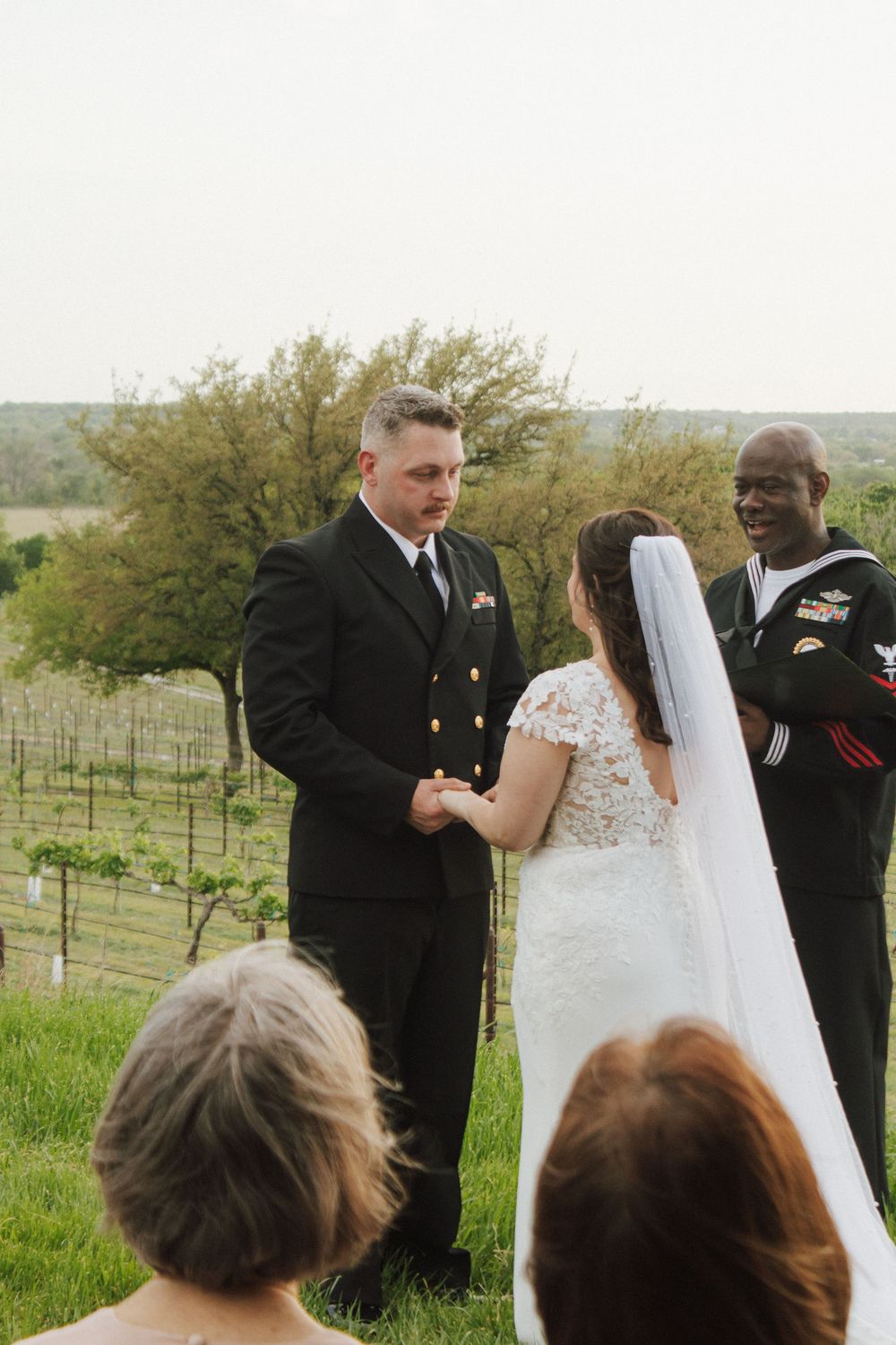 Navy officer groom holds bride's hands during outdoor wedding ceremony at vineyard with officiant smiling nearby.