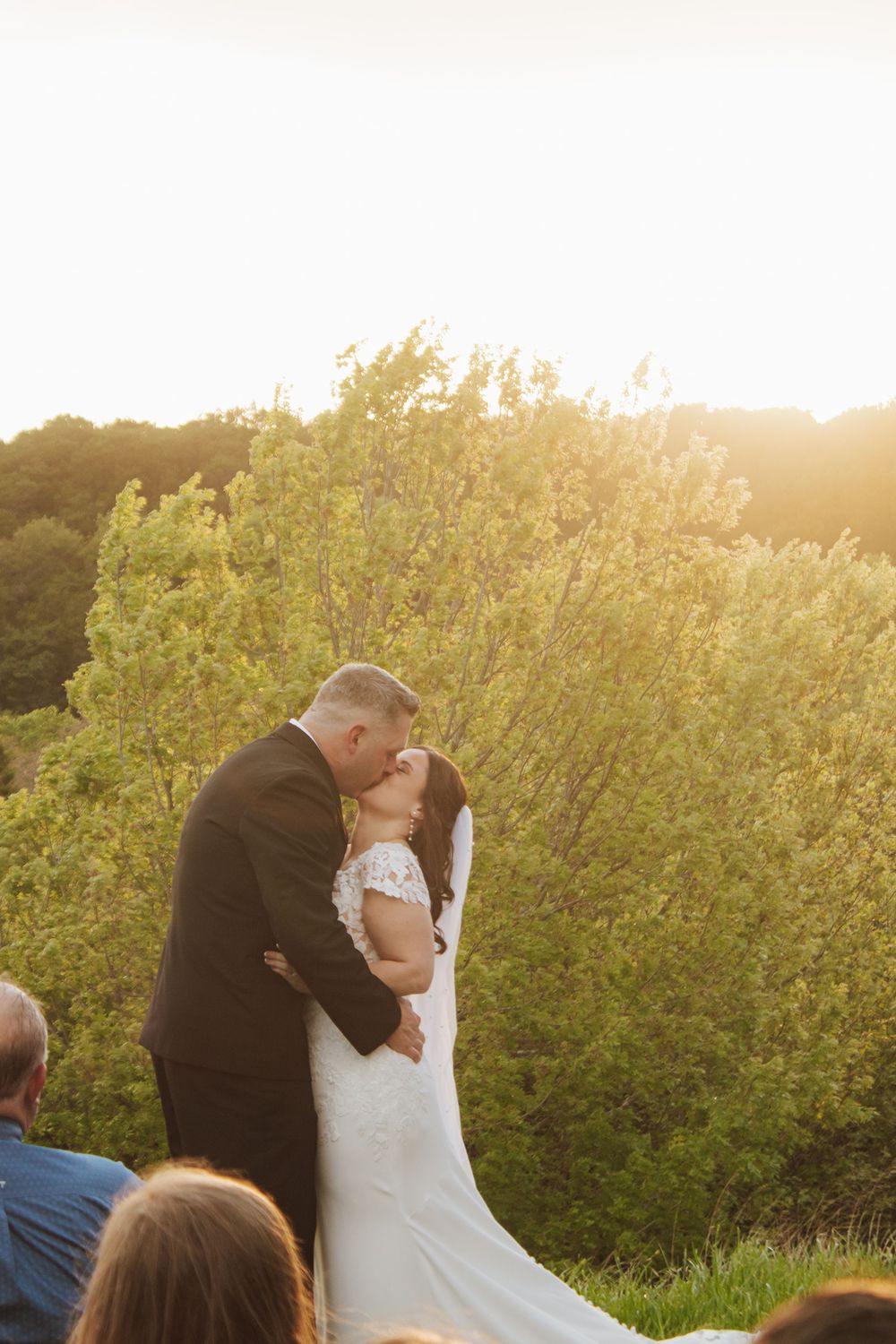 Bride and groom share first kiss at outdoor wedding ceremony with golden sunset light and lush green trees.