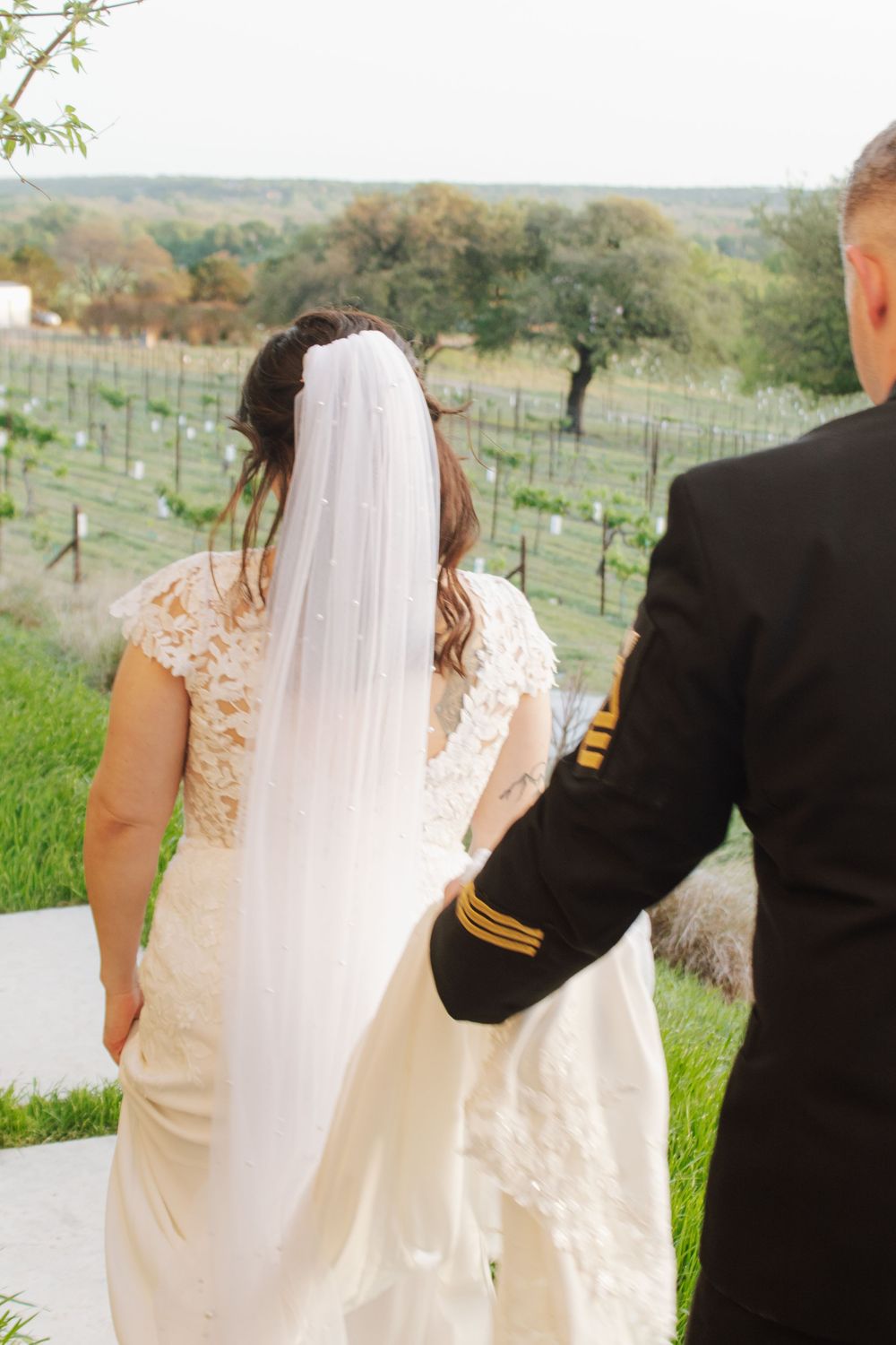 Bride in lace wedding dress and veil walks with groom in military uniform overlooking vineyard.