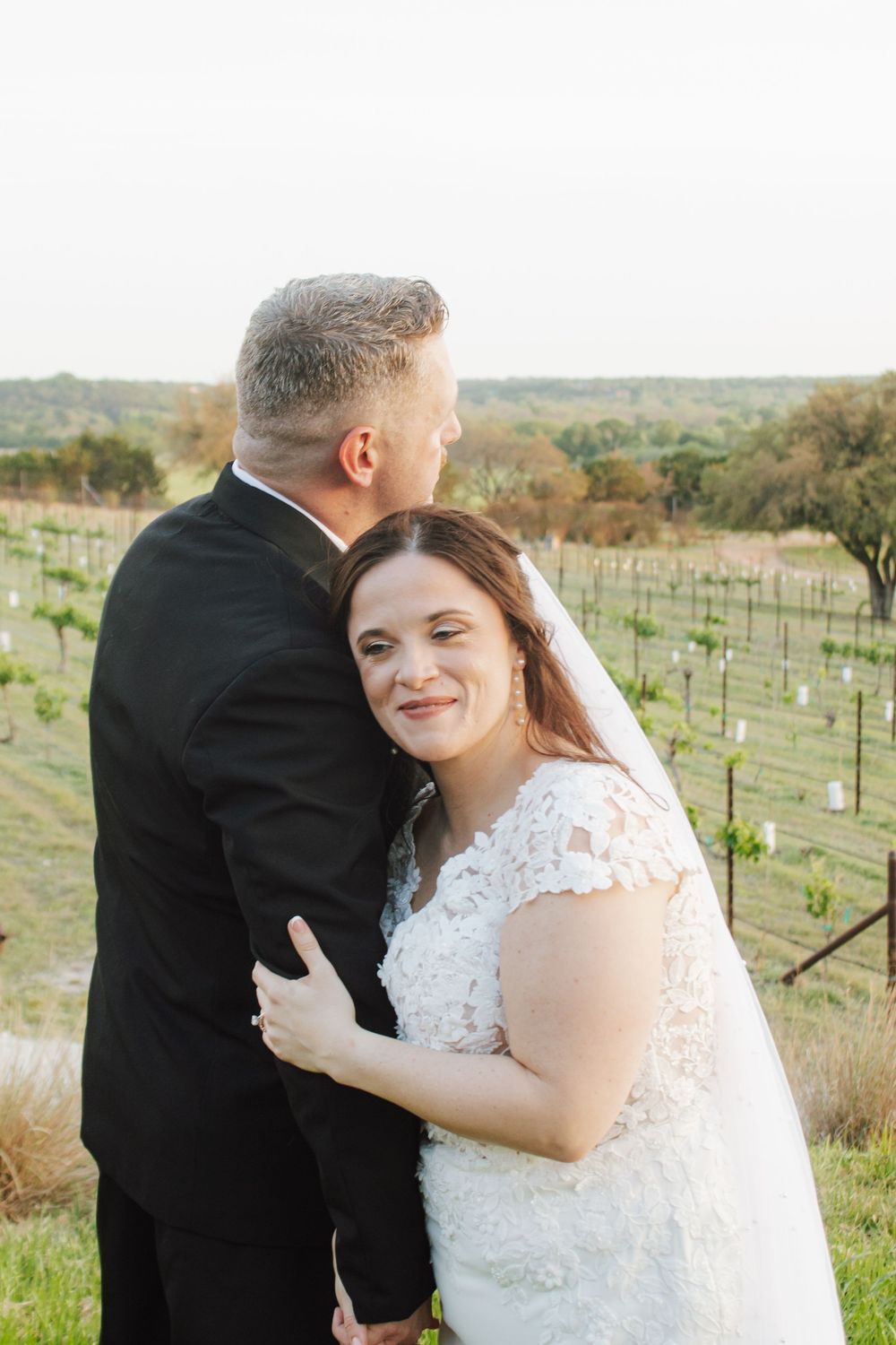 Smiling bride in lace dress embraces groom in black suit at outdoor vineyard wedding venue.