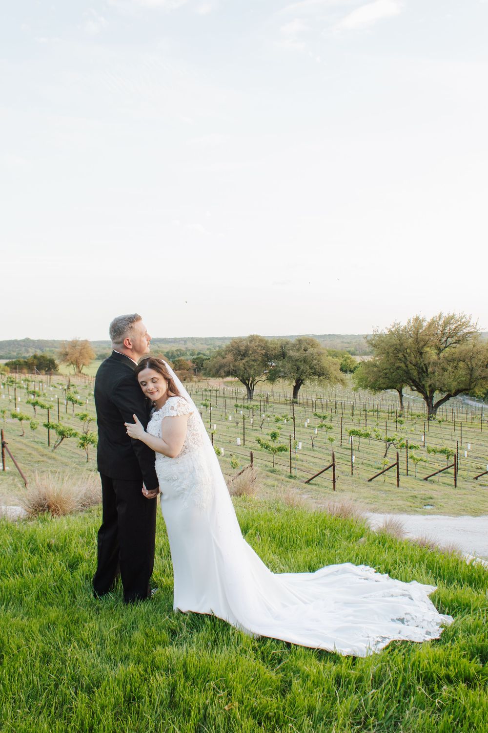 Bride and groom embrace on a lush green hillside overlooking a scenic vineyard on their wedding day.