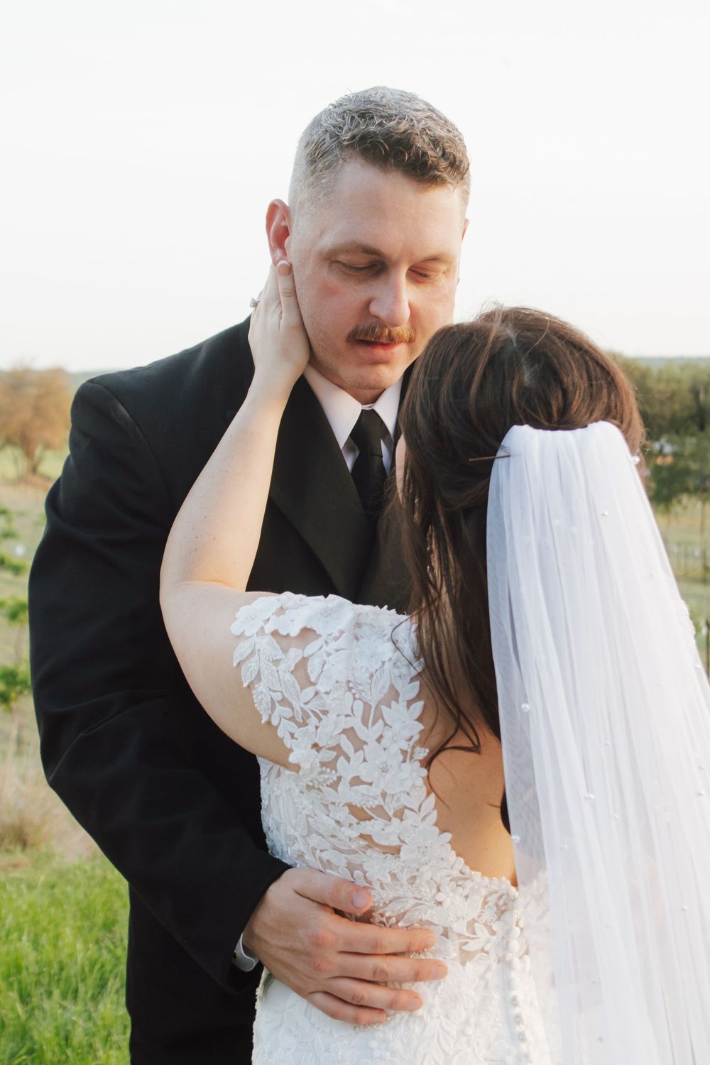 Groom in black suit embraces bride in lace wedding dress outdoors, as she tenderly touches his face.