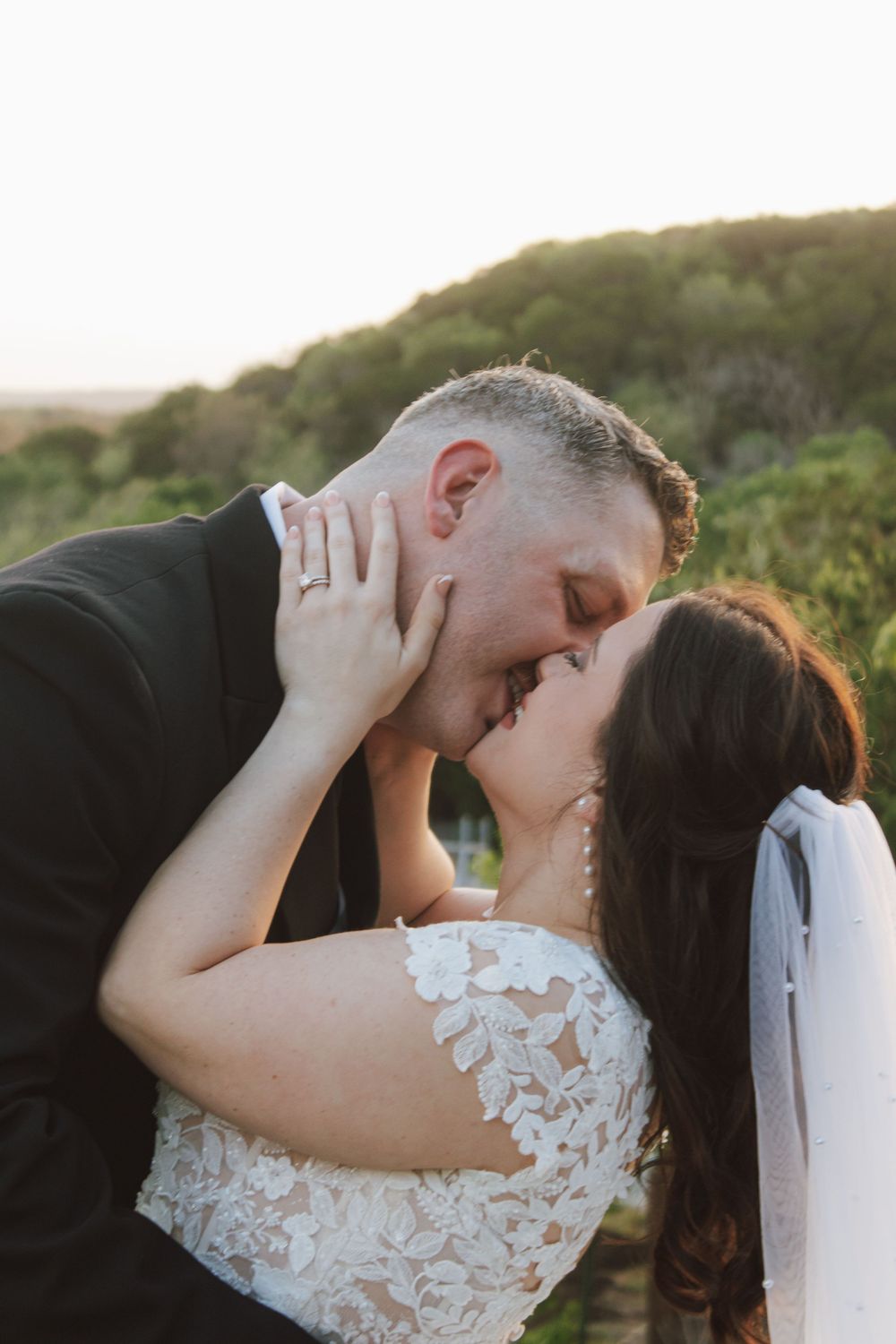 Bride and groom share first kiss at sunset during outdoor wedding ceremony, bride wearing lace dress and veil.