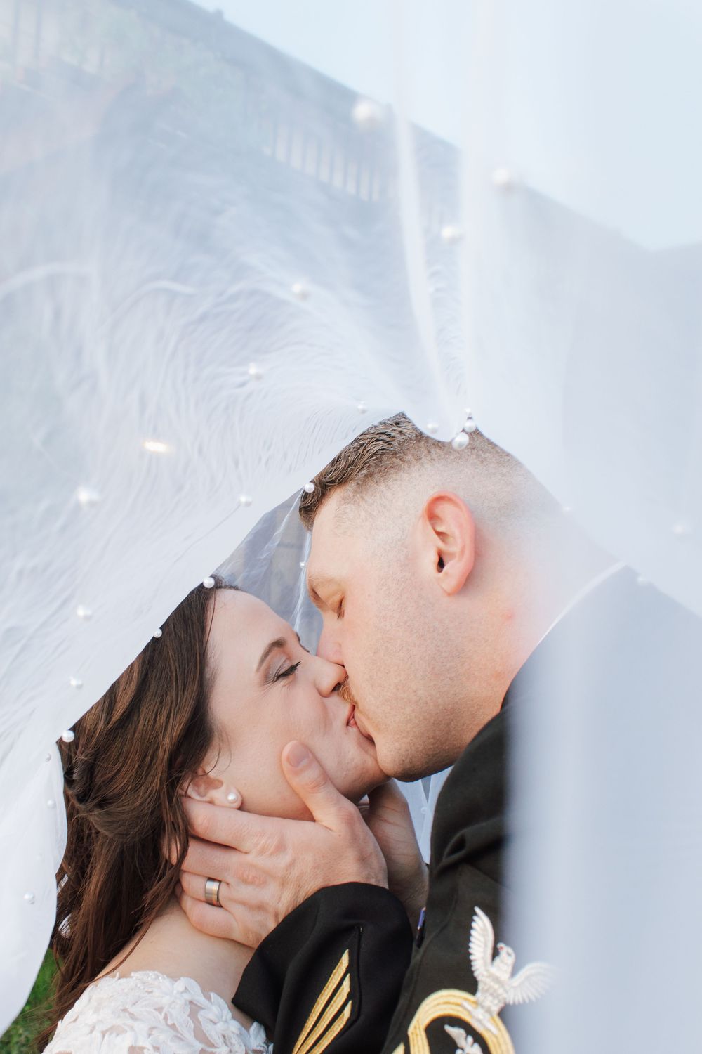 Newlywed couple shares a romantic kiss under a flowing white veil adorned with pearl details.