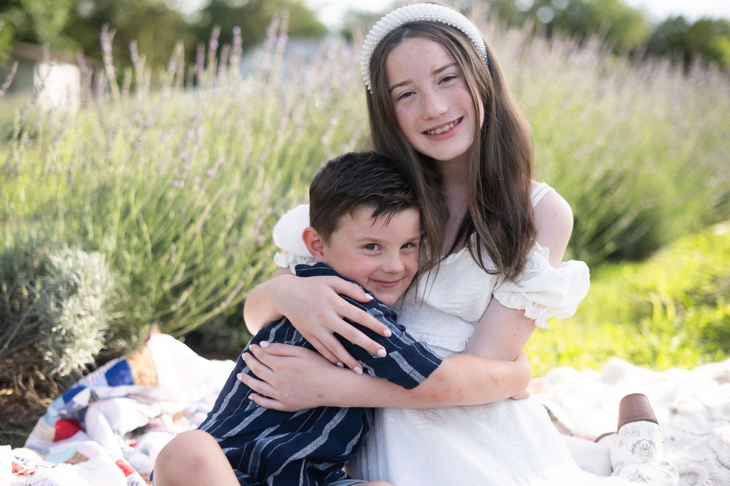 A smiling girl in a white dress hugs a young boy outdoors in a lavender field during a family portrait session.