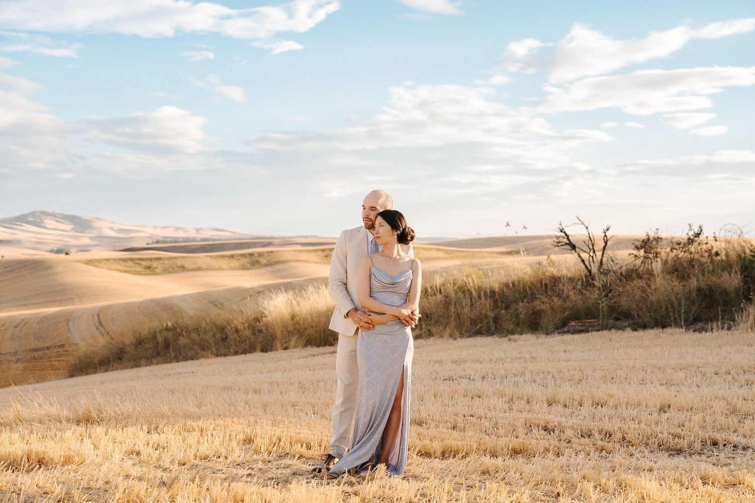 Elegant couple posing in golden Tuscan fields at sunset, man in beige suit, woman in silver gown.