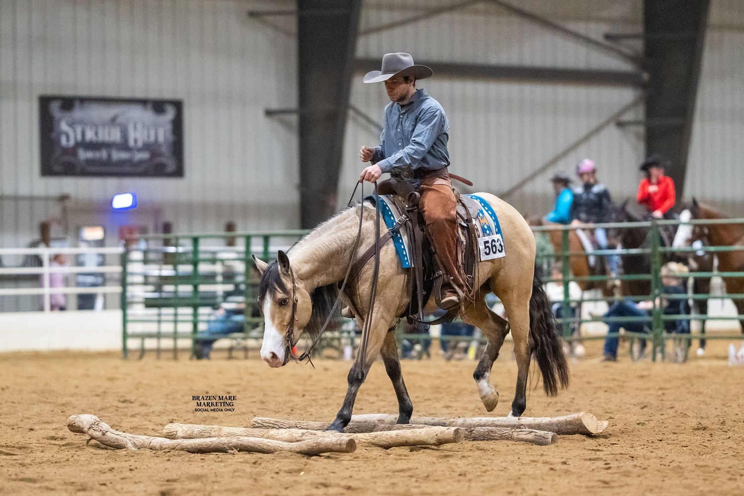 Rider on a buckskin horse navigating a log obstacle in an indoor Western horse show competition.