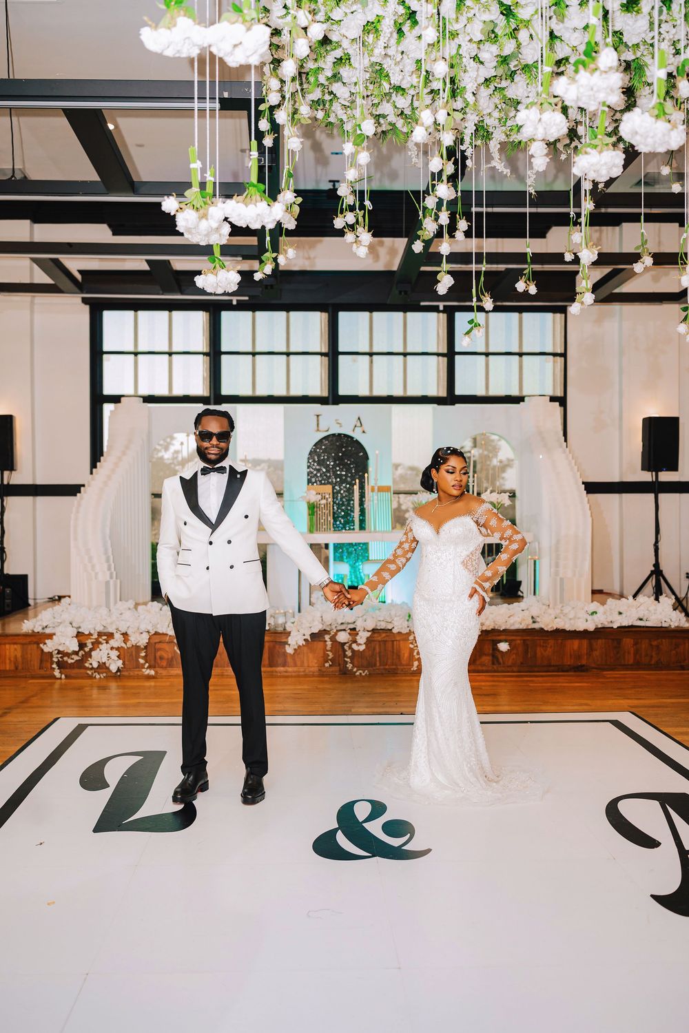Bride and groom hold hands on monogrammed dance floor beneath floral ceiling installation at elegant wedding reception.