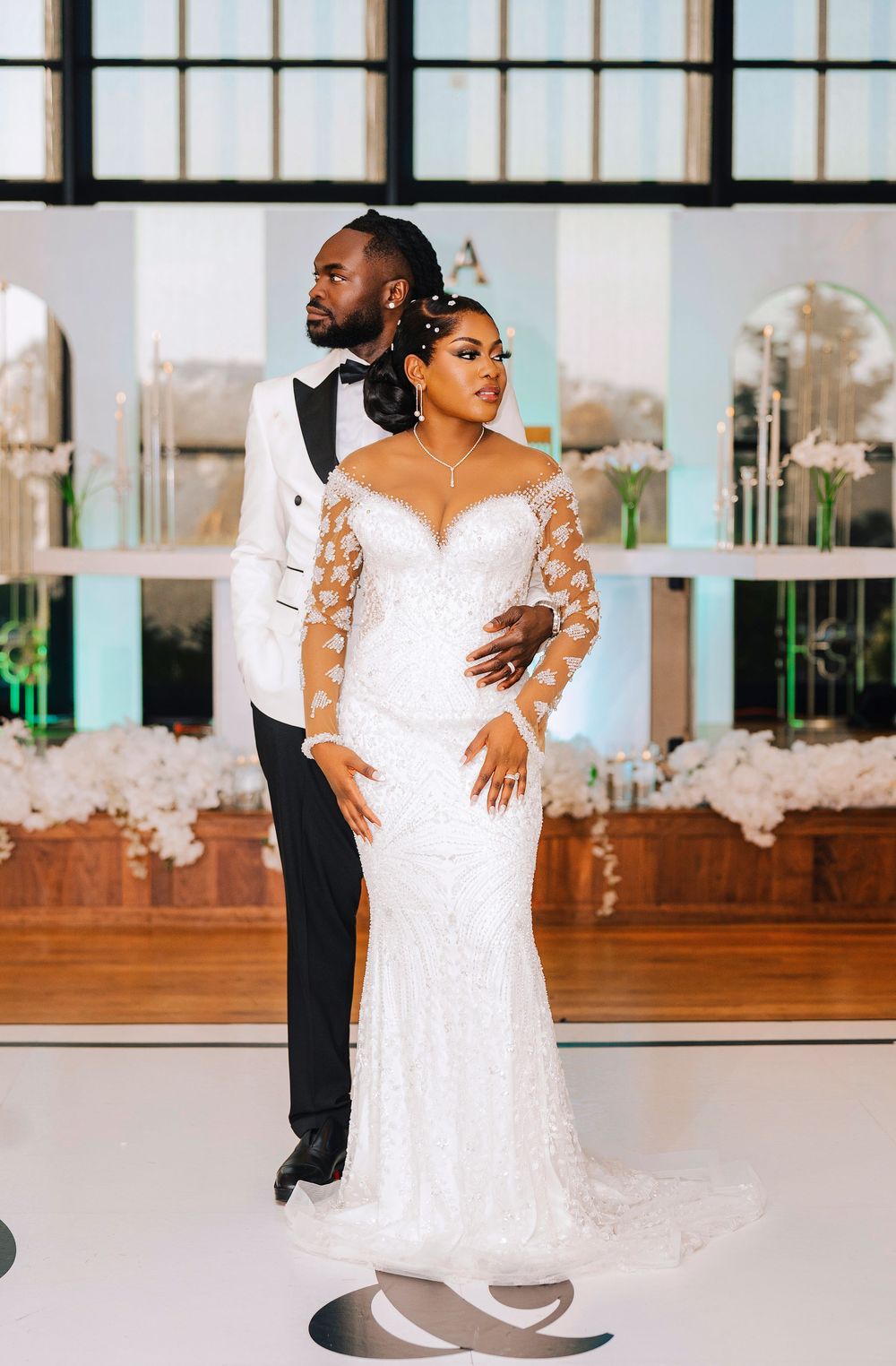 Bride in beaded lace gown and groom in white tuxedo share romantic pose at elegant wedding reception venue.