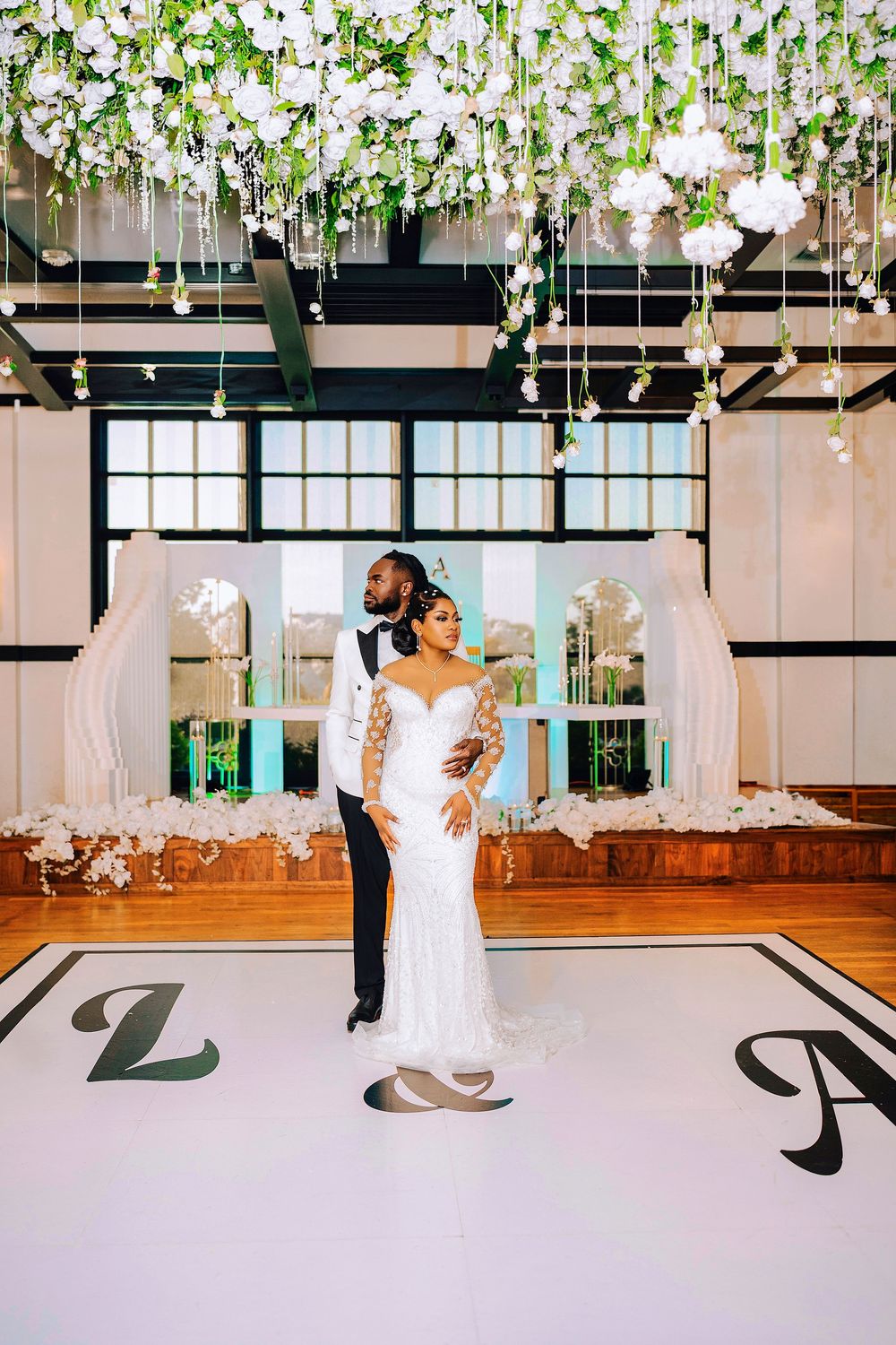 Elegant wedding couple dancing on monogrammed floor beneath hanging floral ceiling installation in decorated ballroom.
