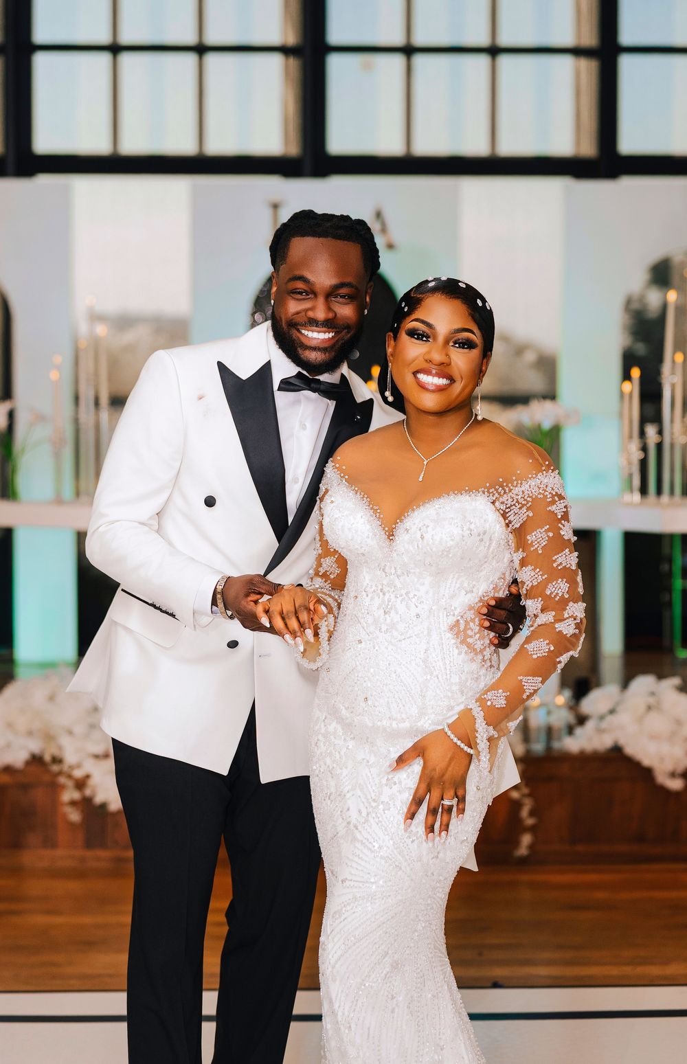 Smiling bride and groom pose together in elegant wedding attire, she in lace gown, he in white tuxedo jacket.