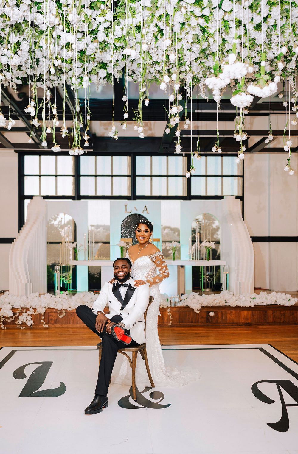 Elegant wedding couple poses on dance floor under floral ceiling installation with white roses and greenery.