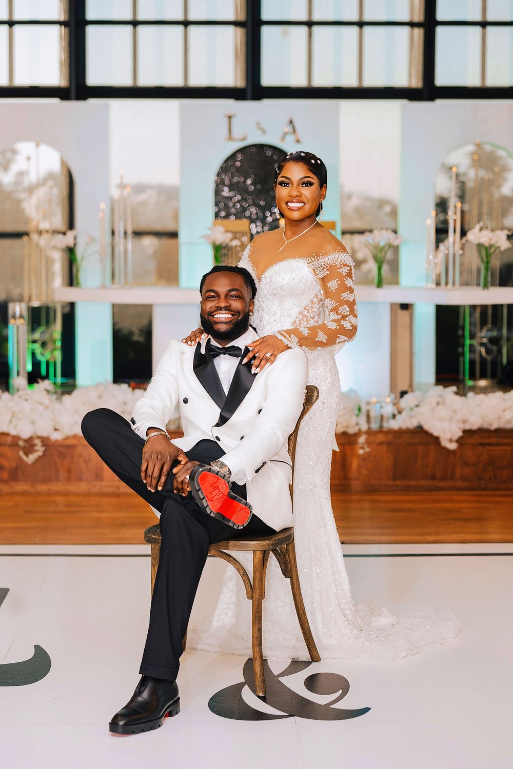 Bride in lace gown stands behind groom in white tuxedo seated on chair at elegant wedding reception.