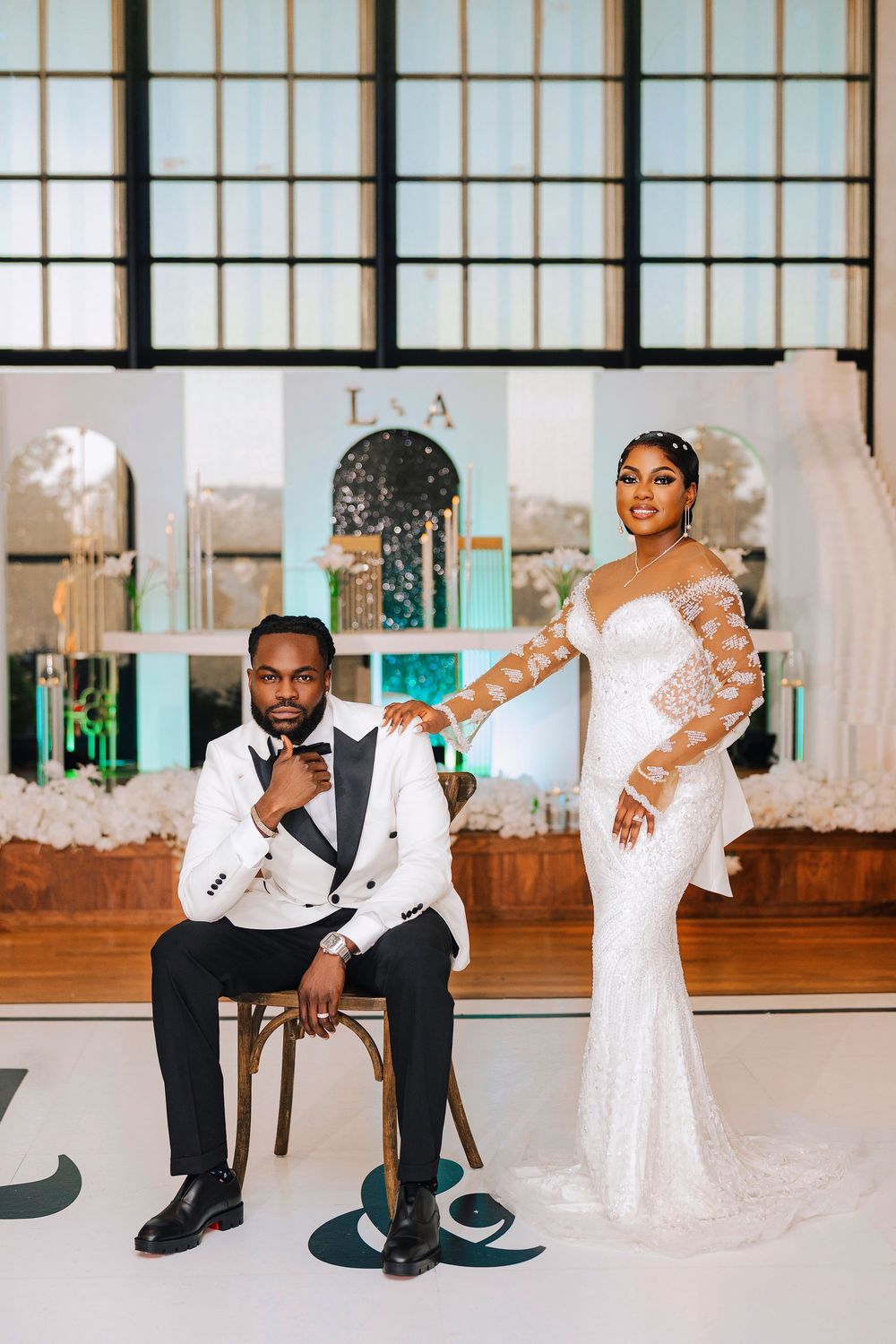 Elegant bride in lace gown and groom in white tuxedo posing at a teal-accented wedding reception venue.