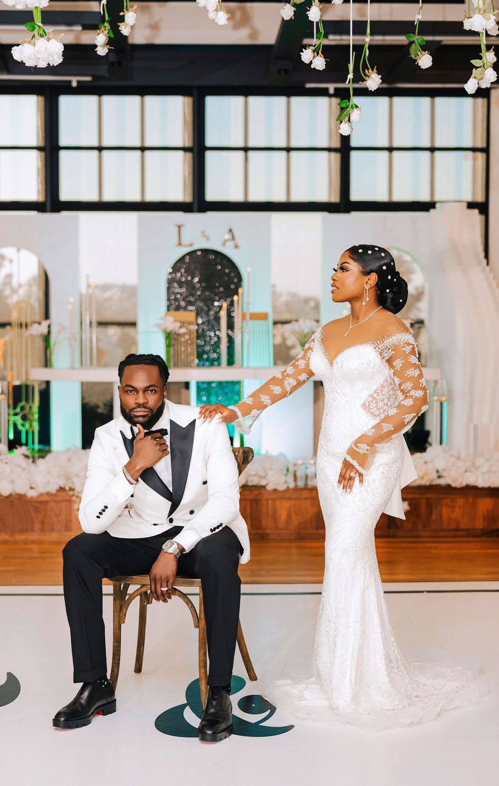 Elegant bride and groom in white wedding attire pose in a decorated venue with floral arrangements.