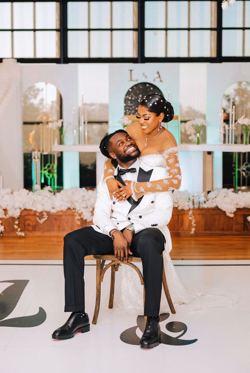 Bride embraces groom seated on wooden chair on monogrammed dance floor at elegant white floral wedding reception.