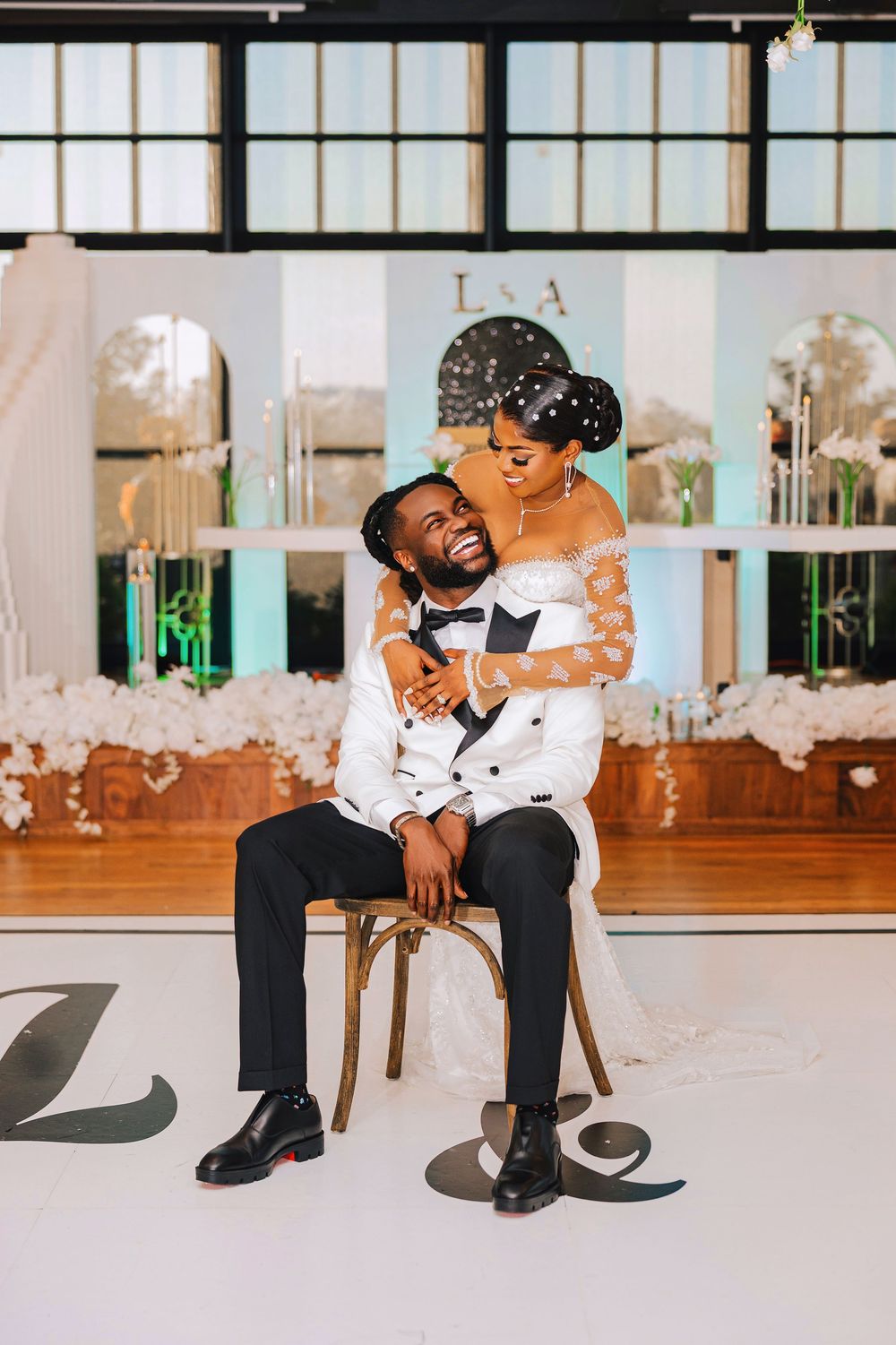 Joyful Black bride and groom share a tender moment on wedding dance floor, groom seated as bride embraces him.