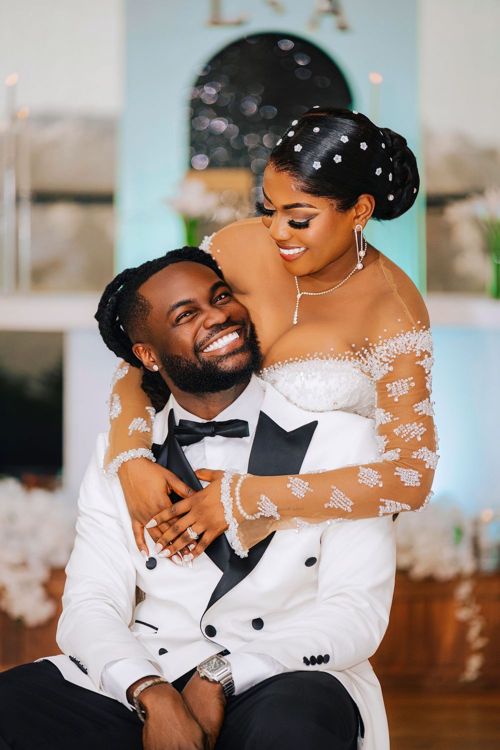 Groom in white tuxedo smiles as bride in lace wedding dress embraces him from behind at wedding reception.