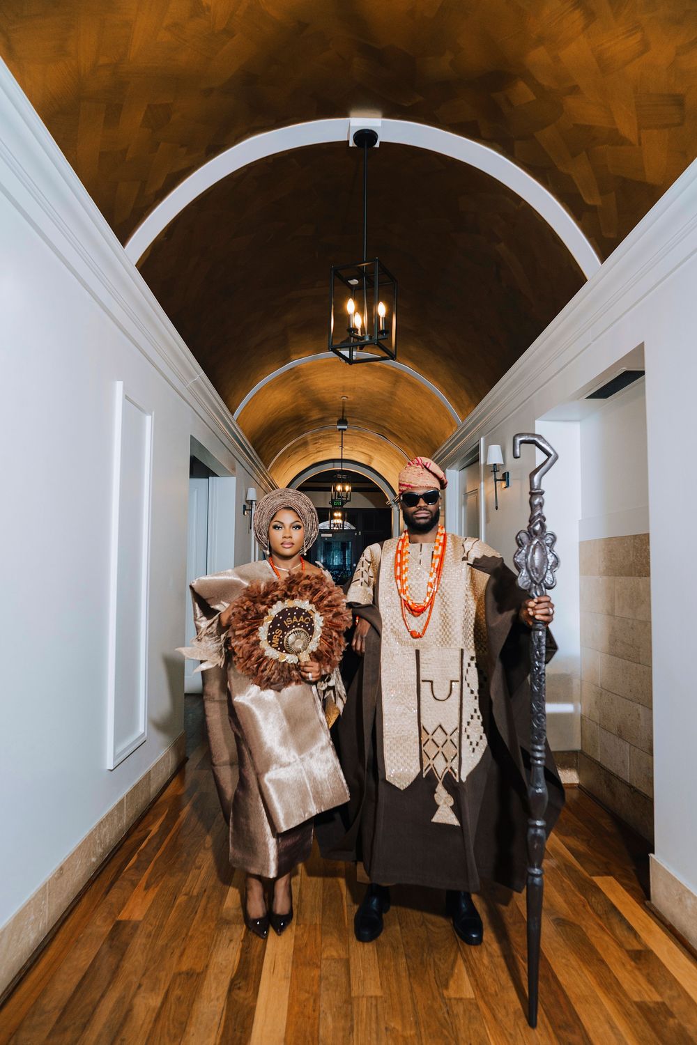 Couple in traditional African attire poses in elegant arched hallway, man holding ceremonial staff.