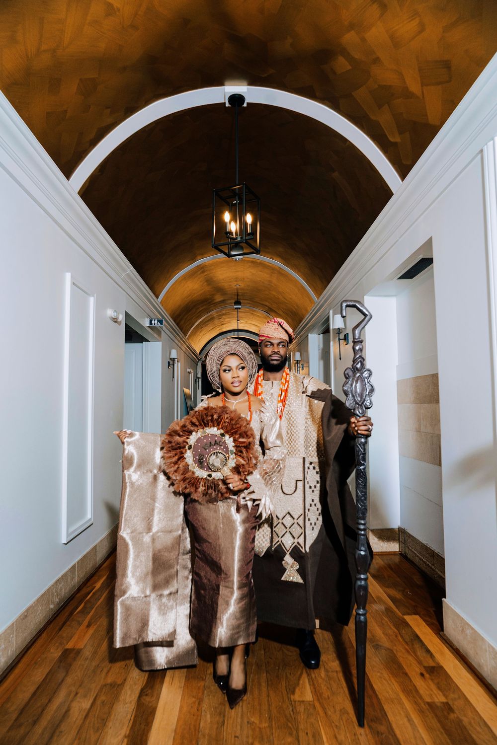 Couple in traditional Nigerian wedding attire pose in an arched hallway, groom holding a ceremonial staff.