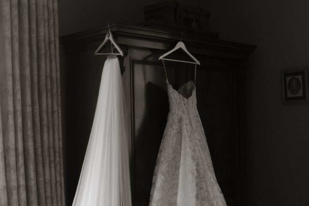 Black and white photo of a lace wedding dress and veil hanging on hangers in front of a dark wooden armoire.
