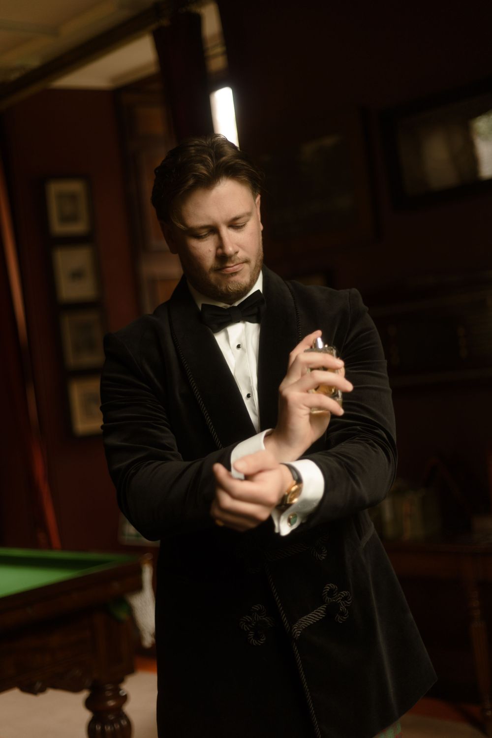 Elegant man in black velvet tuxedo adjusting cufflinks in a sophisticated billiard room setting.