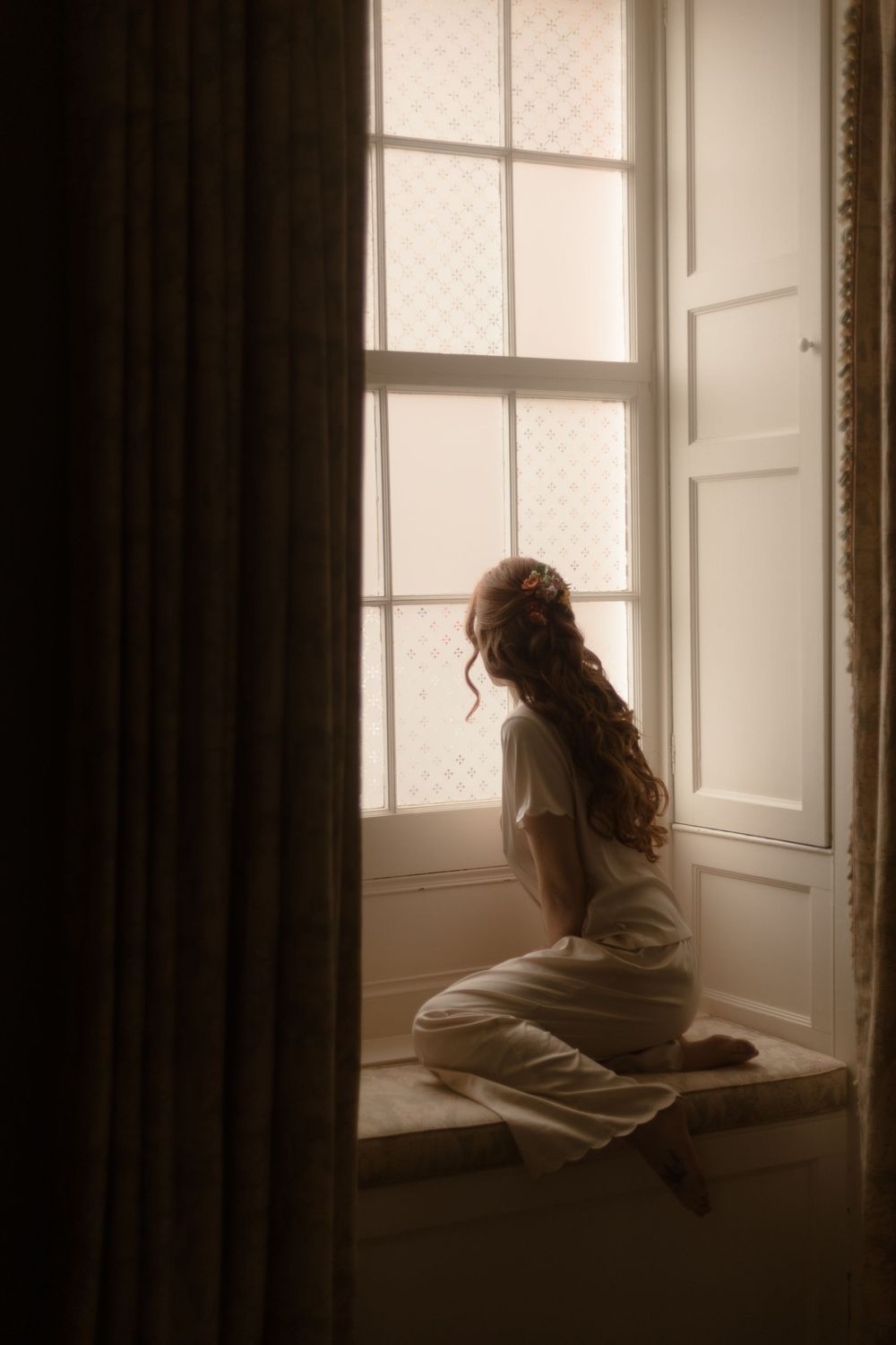 A bride in white sits on a sunlit window seat, her long curly hair adorned with a floral piece, gazing outside.