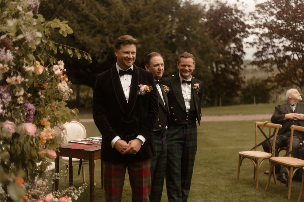 Three men in formal Scottish attire smile at an outdoor wedding ceremony surrounded by floral arrangements.