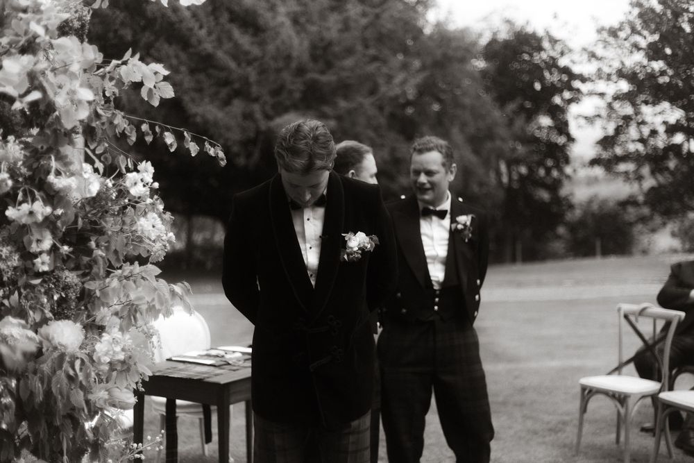 Two grooms in tuxedos share a tender moment at an outdoor wedding ceremony surrounded by floral arrangements.