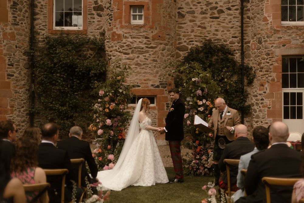 Bride and groom exchange vows outdoors at a rustic stone castle wedding ceremony with floral arch and seated guests.