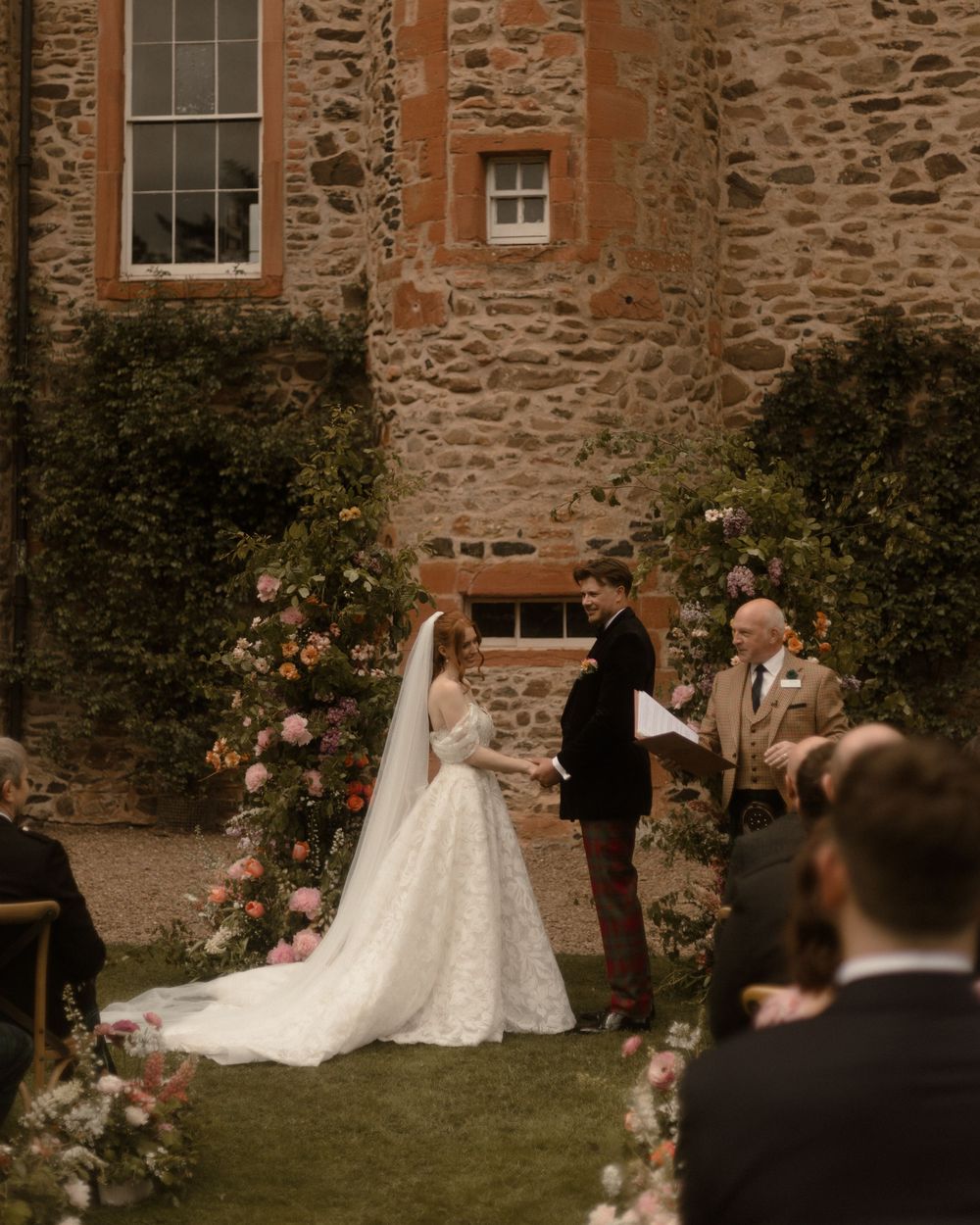 Outdoor wedding ceremony at a stone castle venue with bride in lace gown and floral arch backdrop.