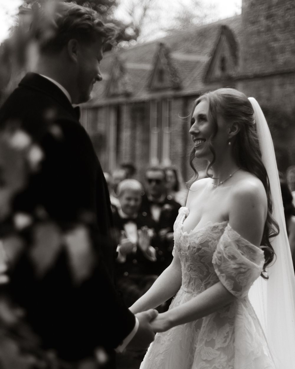 Black and white photo of smiling bride in lace gown holding hands with groom during outdoor wedding ceremony.