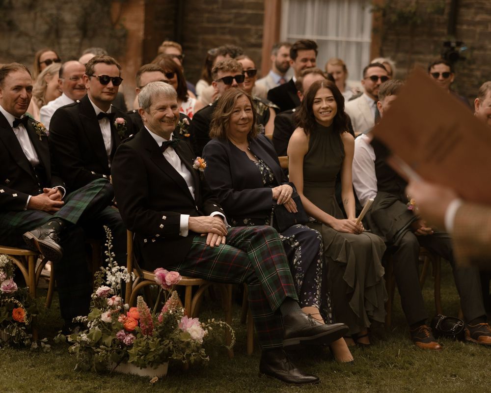 Wedding guests seated outdoors smiling during a ceremony, dressed in formal attire including kilts, at a rustic venue.