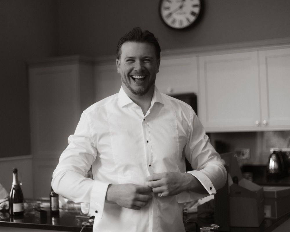 Smiling man in white dress shirt buttoning cuffs in a kitchen, black and white photo.