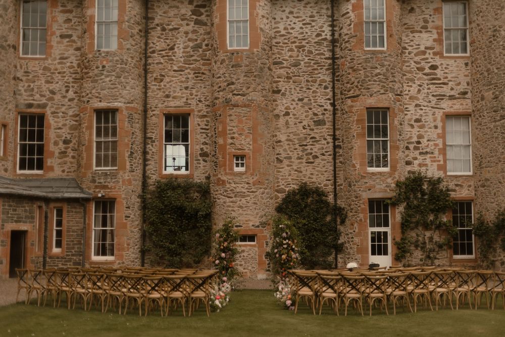 Outdoor wedding ceremony setup with wooden chairs on lawn before a historic stone castle venue.