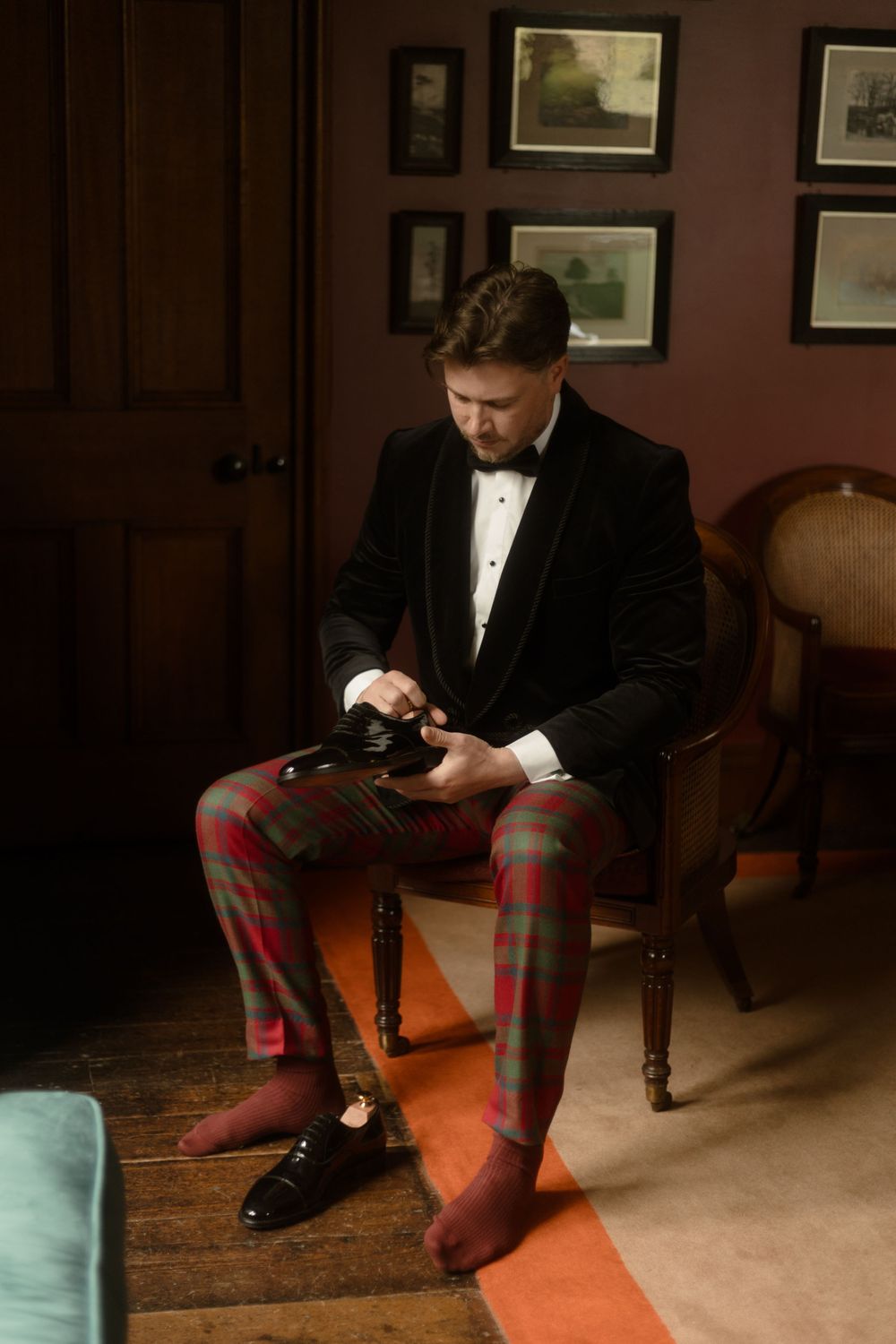 Groom in velvet tuxedo jacket and plaid trousers sits in chair adjusting his shoe before wedding ceremony.