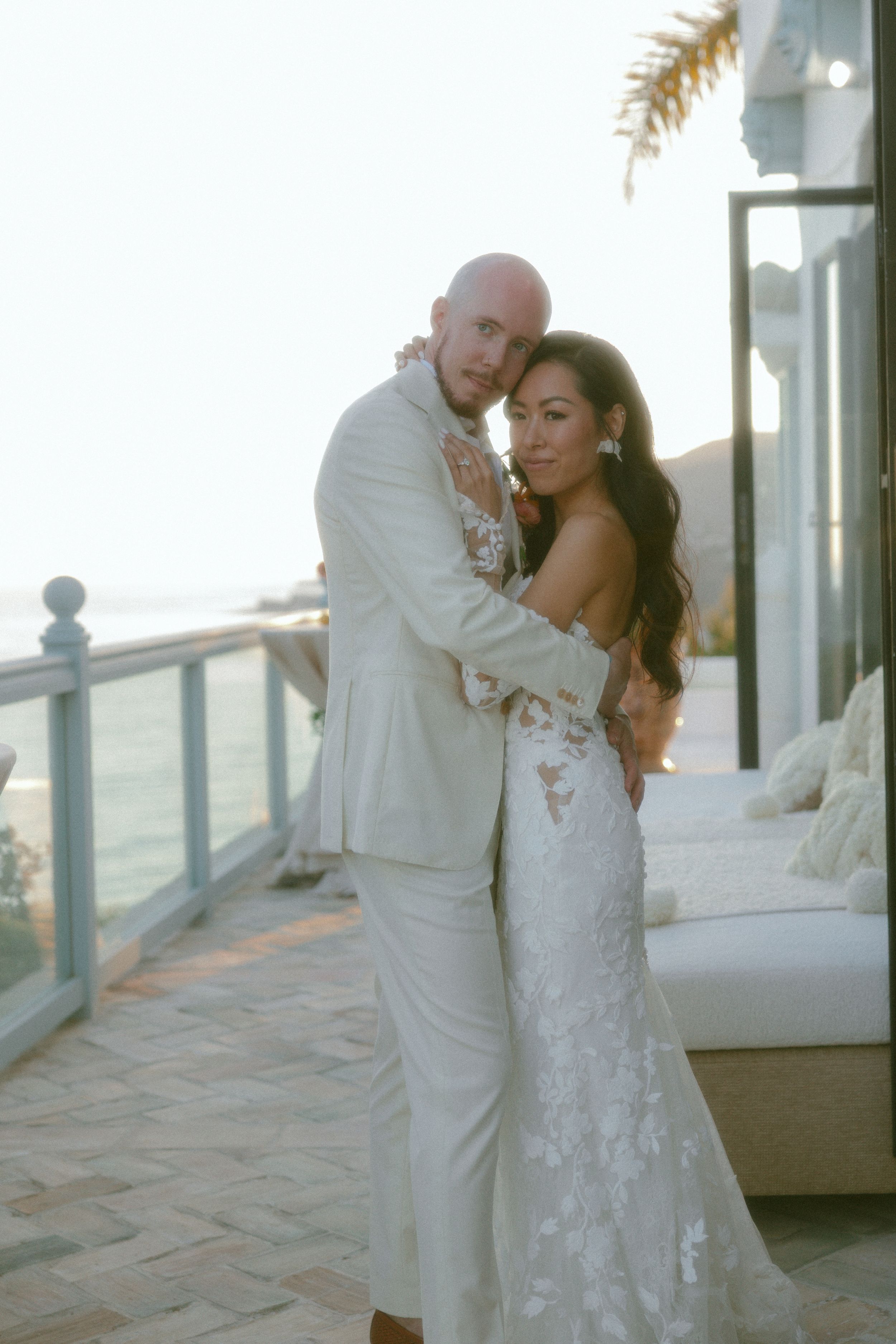 Couple embracing on a beachfront balcony, groom in white suit and bride in lace wedding gown at sunset.