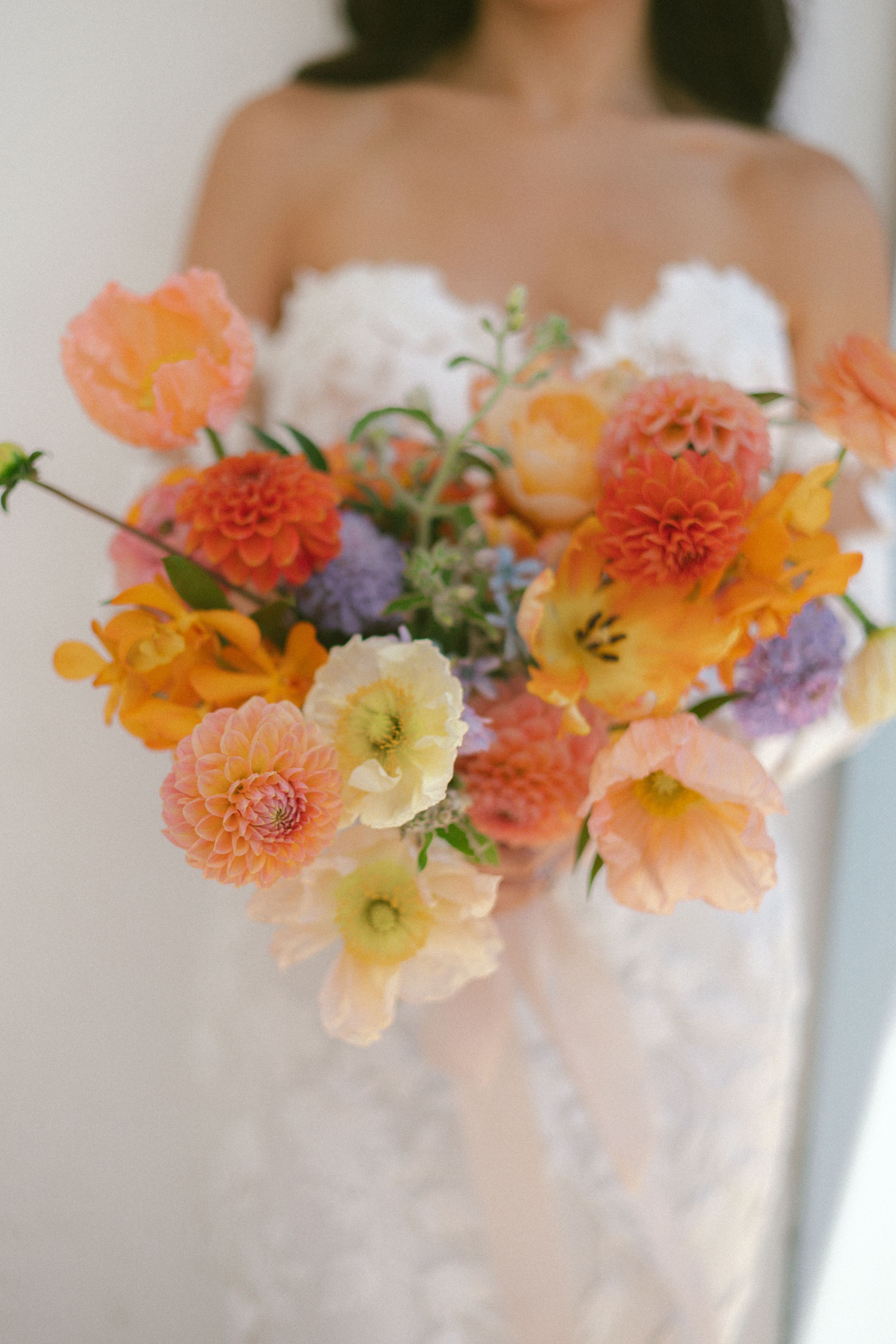 Bride holding a vibrant wedding bouquet with orange dahlias, pink poppies, yellow tulips, and purple blooms in white dress.