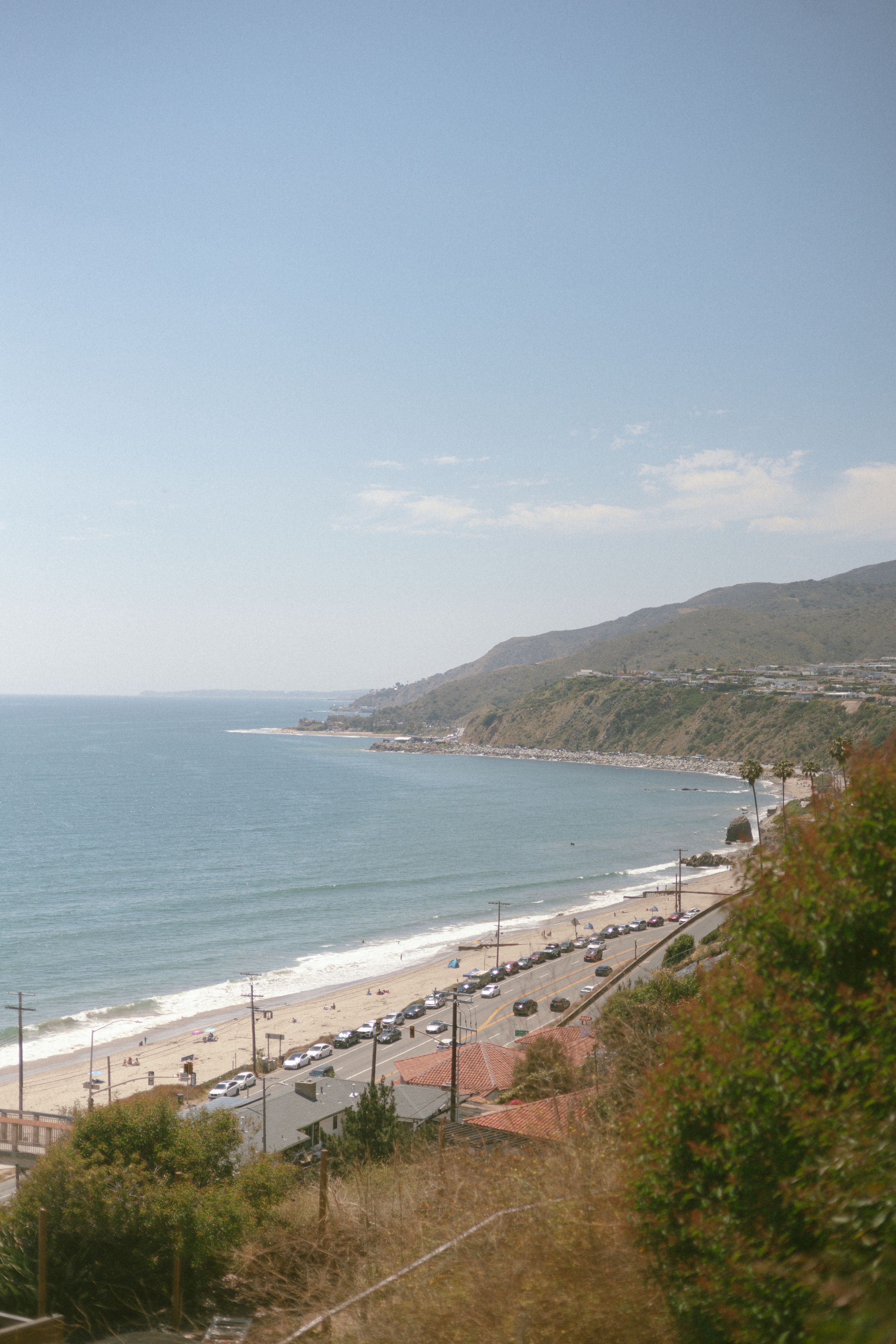 Aerial view of a scenic coastal highway along a sandy beach with parked cars, rolling hills, and calm blue ocean waters.