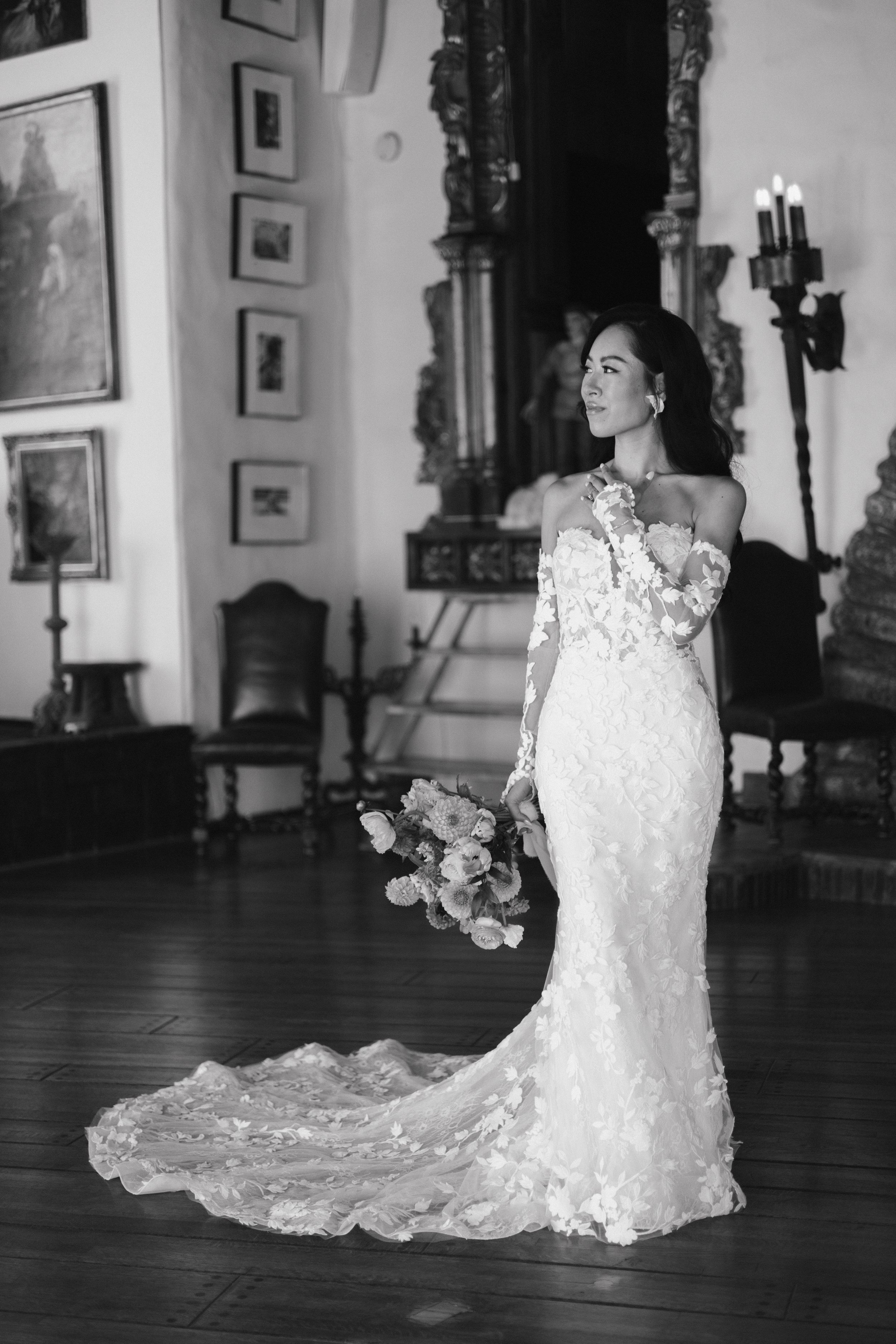 Black and white photo of a bride in a lace off-shoulder gown with train, holding a bouquet in an elegant hall.