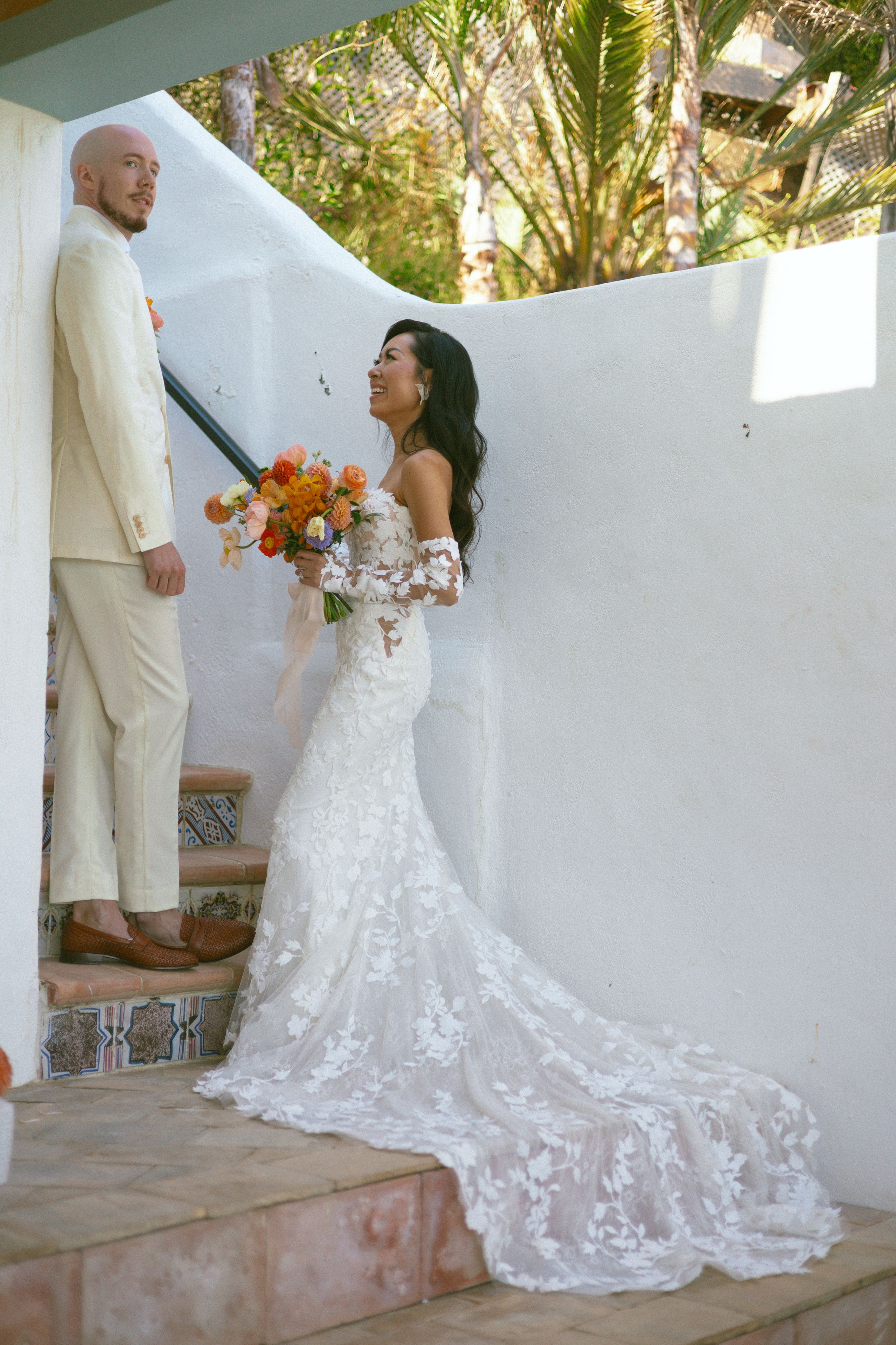 Bride in lace mermaid gown with floral bouquet stands beside groom in tan suit on tiled steps at outdoor wedding.