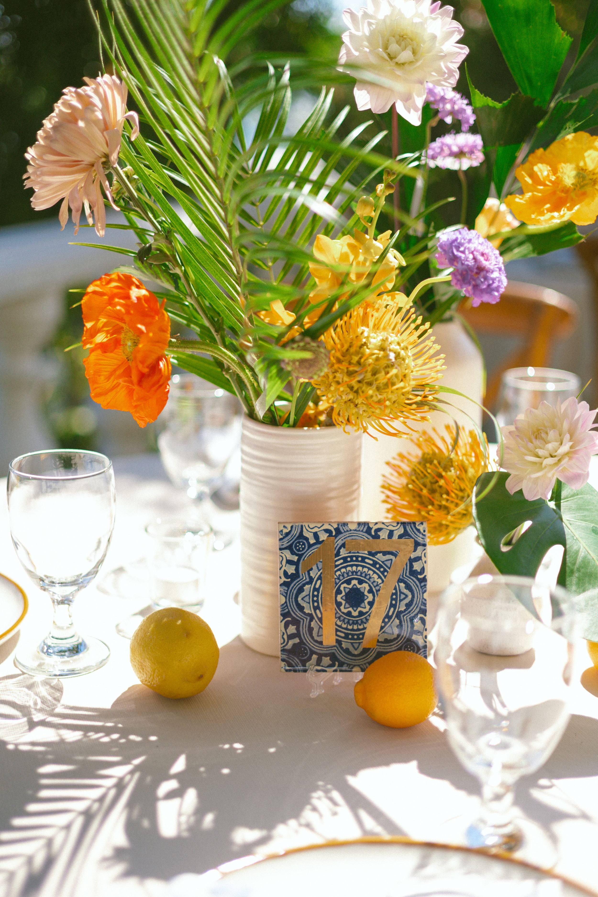 Colorful tropical wedding table centerpiece with orange, yellow, and pink flowers, palm leaves, lemons, and a blue tile table number.
