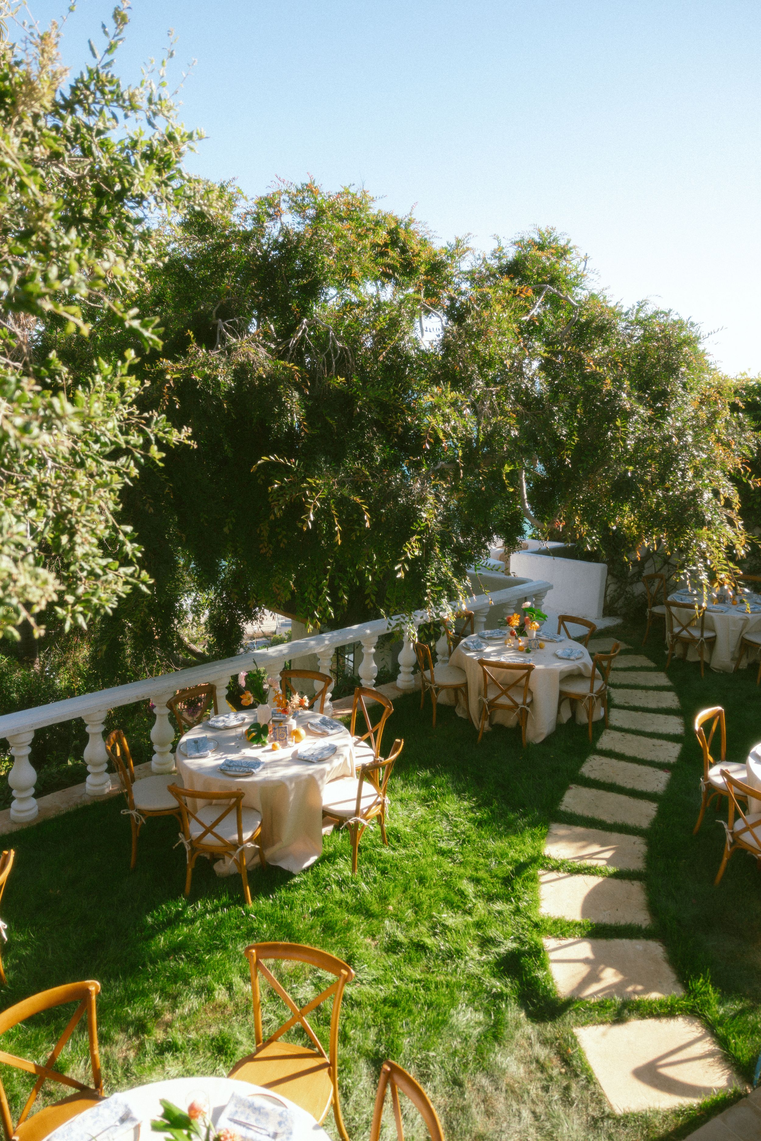 Elegant outdoor garden dining setup with round tables, wooden cross-back chairs, lush greenery, and a stone pathway on green lawn.
