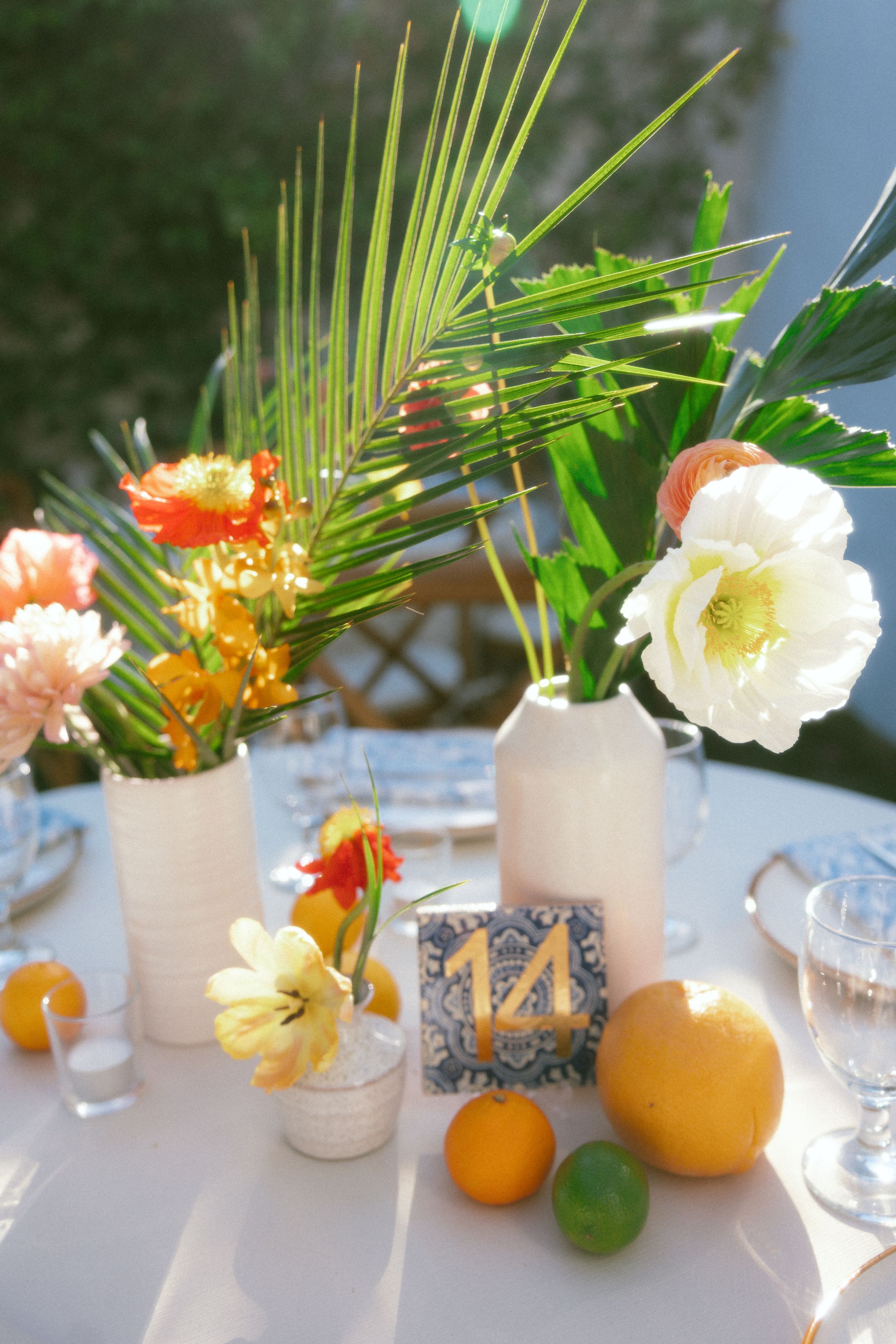 Tropical wedding table centerpiece with white vases, colorful flowers, palm leaves, and citrus fruits on table 14.