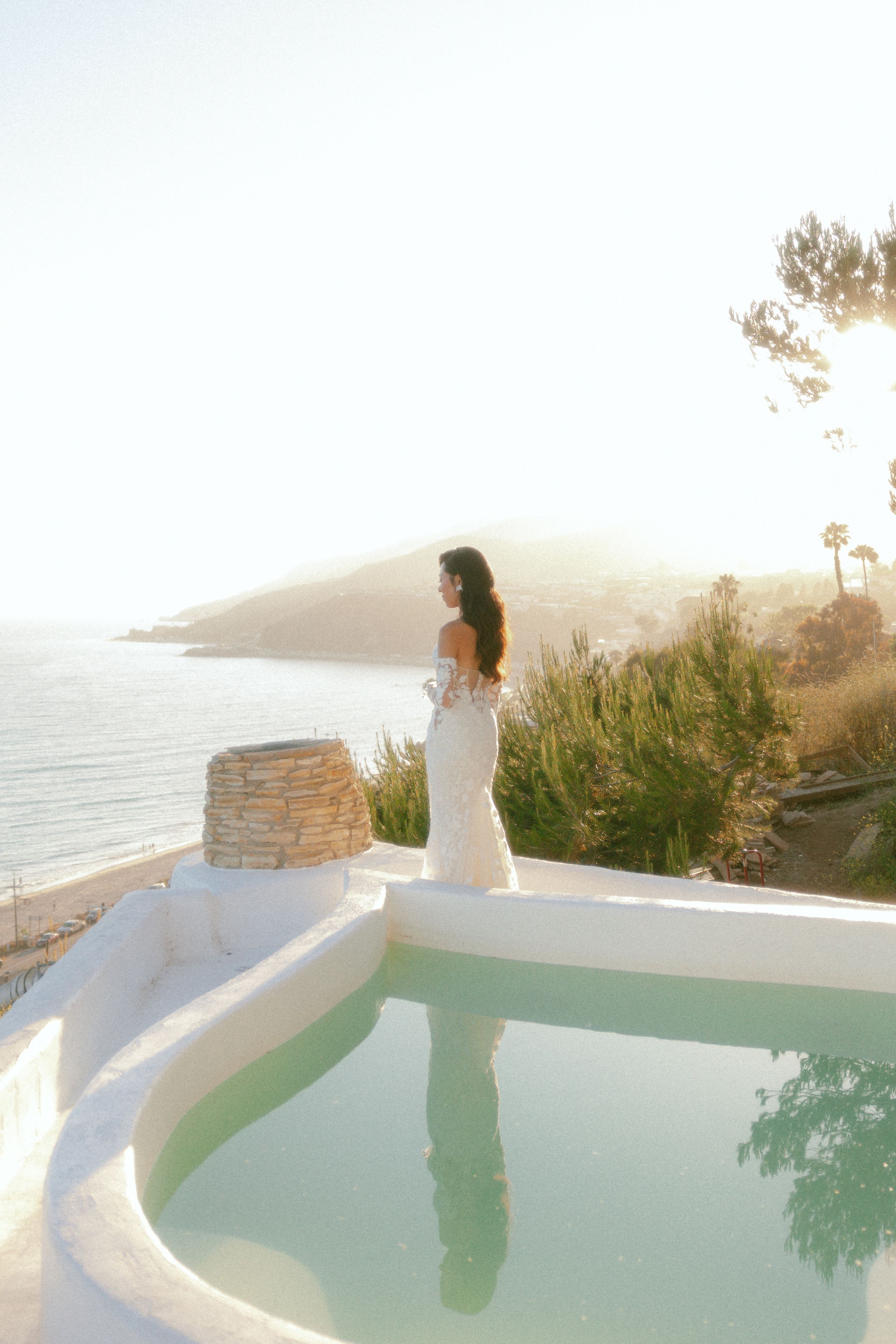 Bride in white gown standing by a pool with ocean and hillside views in soft golden light.