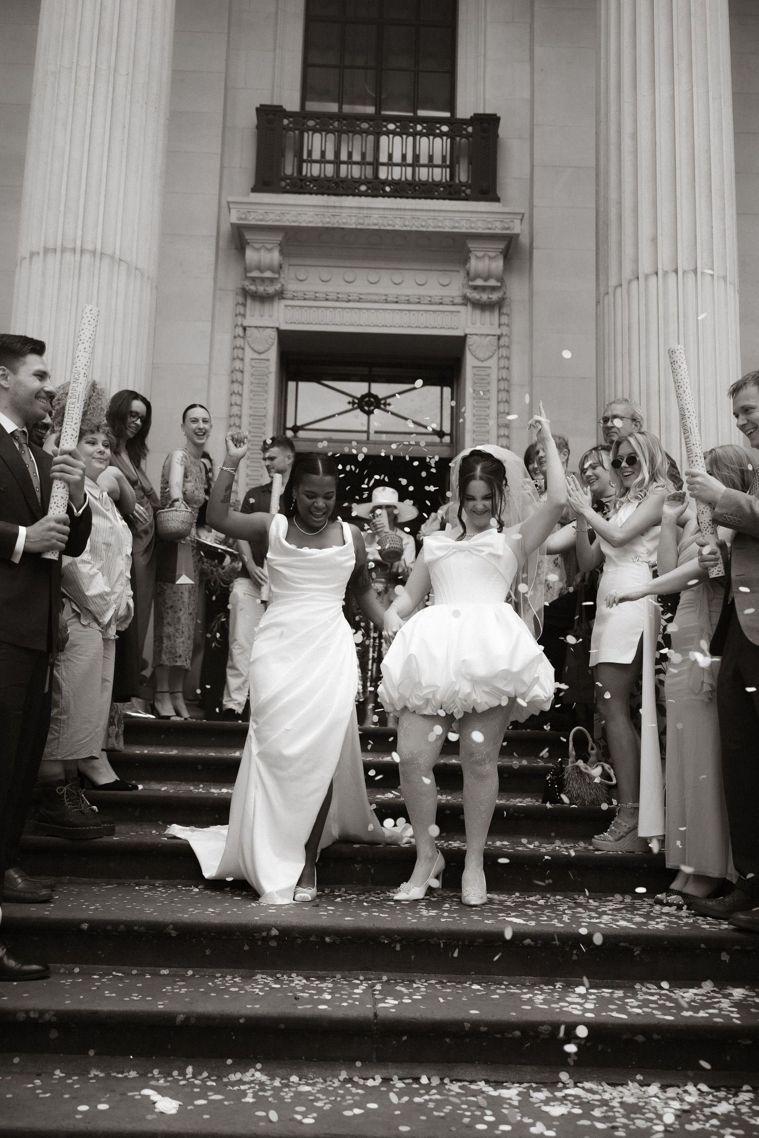 A cheerful wedding party celebrates on the grounds of a historic stone manor house in muted sepia tones.