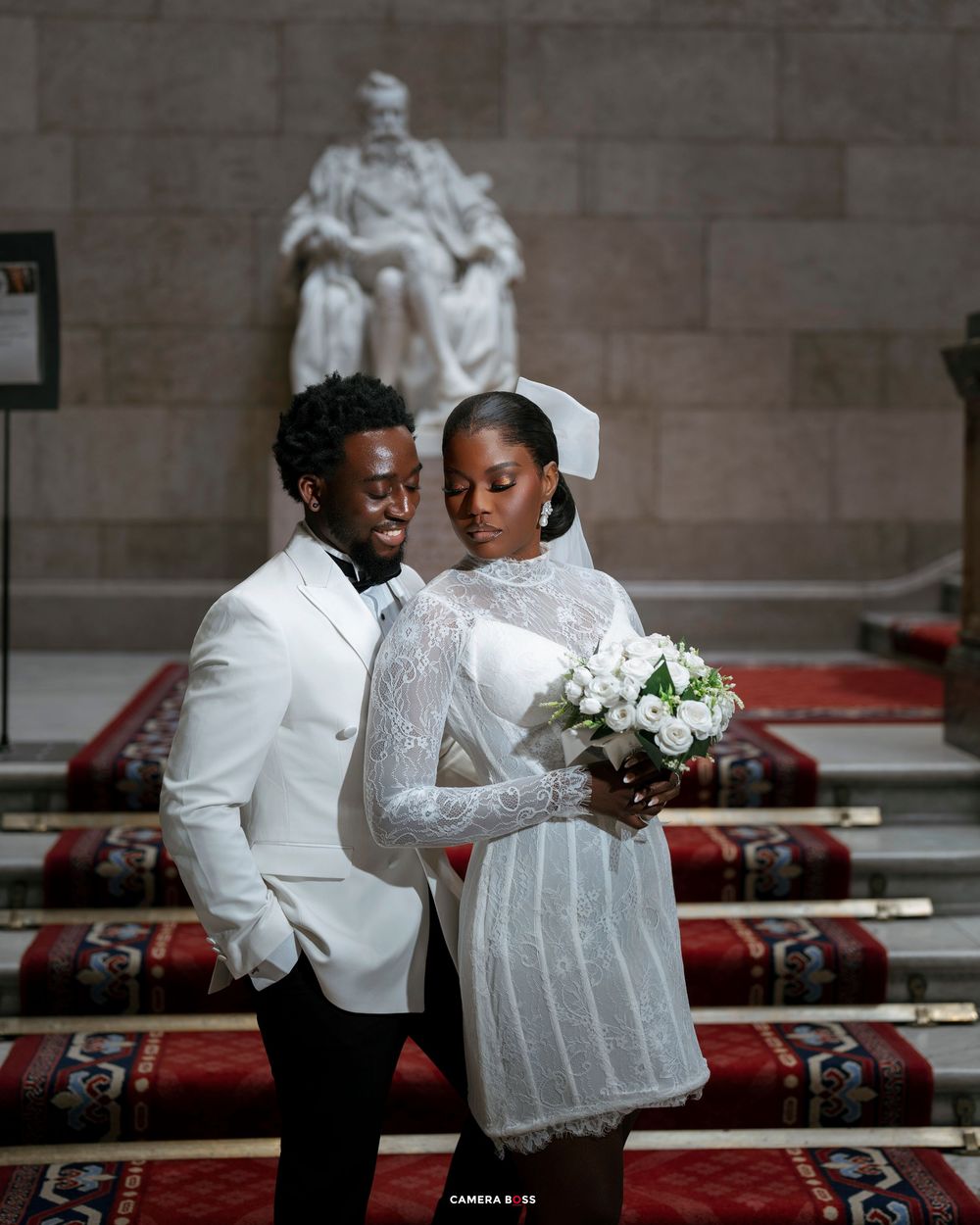 Elegant couple in white wedding attire pose before a grand statue in a formal hall, bride holding white bouquet.