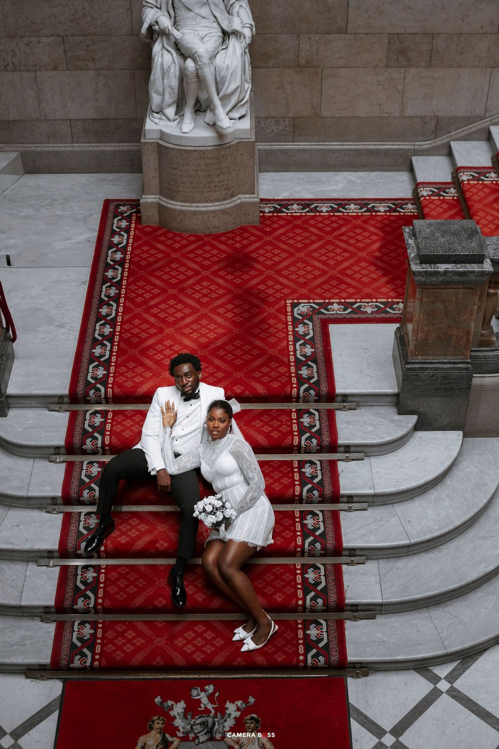 Newlywed couple in wedding attire sitting on red carpeted stairs in elegant hall with marble statue backdrop.
